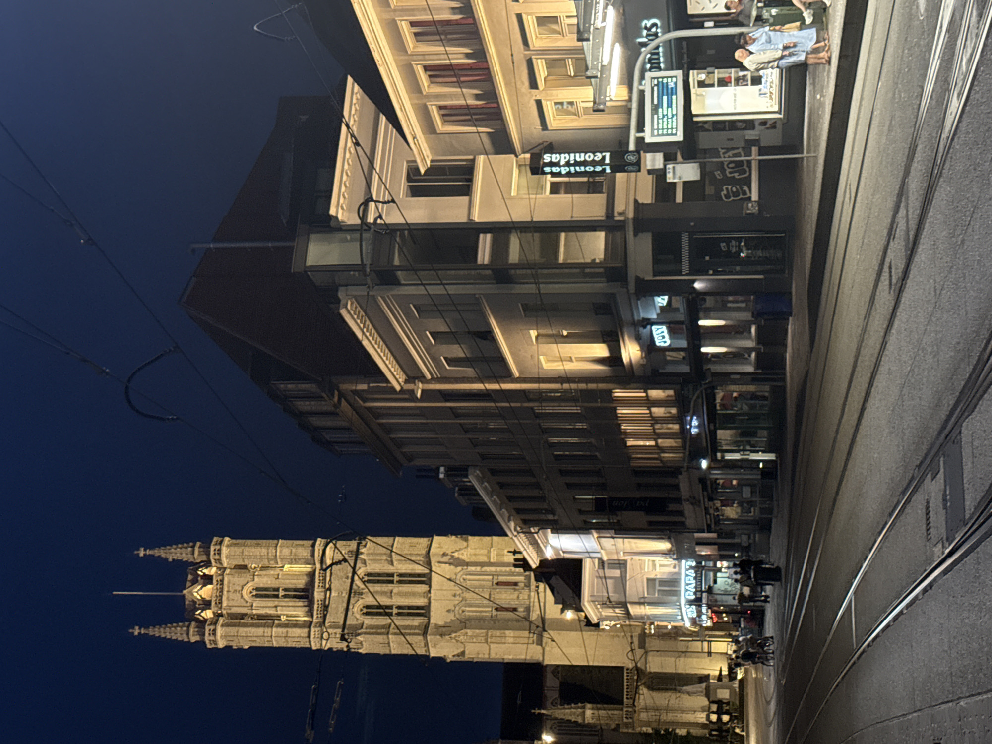 The image captures a nighttime street scene in Ghent, Belgium, focusing on the Sint-Niklaaskerk (Saint Nicholas' Church) tower and surrounding buildings. The church tower, illuminated by artificial lights, is a prominent feature on the left side of the frame, exhibiting Gothic architectural style with spires and arched windows. Adjacent to the tower, a series of buildings line the street, displaying various architectural elements and illuminated windows.
In the foreground, tram tracks run along the asphalt road, indicating active public transportation. Several streetlights and overhead wires for the tram system are visible, contributing to the urban nighttime ambiance.
On the sidewalk, several individuals are present. A few people are standing near a bus stop or information display, which features text and numbers, possibly indicating public transport schedules. One person appears to be a woman in a light blue dress and another is a man in a light-colored jacket and dark pants. Further down the street, a group of people are gathered near a building entrance, and some individuals are on bicycles.
Businesses with illuminated signs, such as "JOEY" and "Leonidas," are visible along the street, suggesting commercial activity. The overall atmosphere is one of a vibrant, well-lit European city at night, with a blend of historical architecture and modern urban life. The sky is dark, indicating late evening or night.