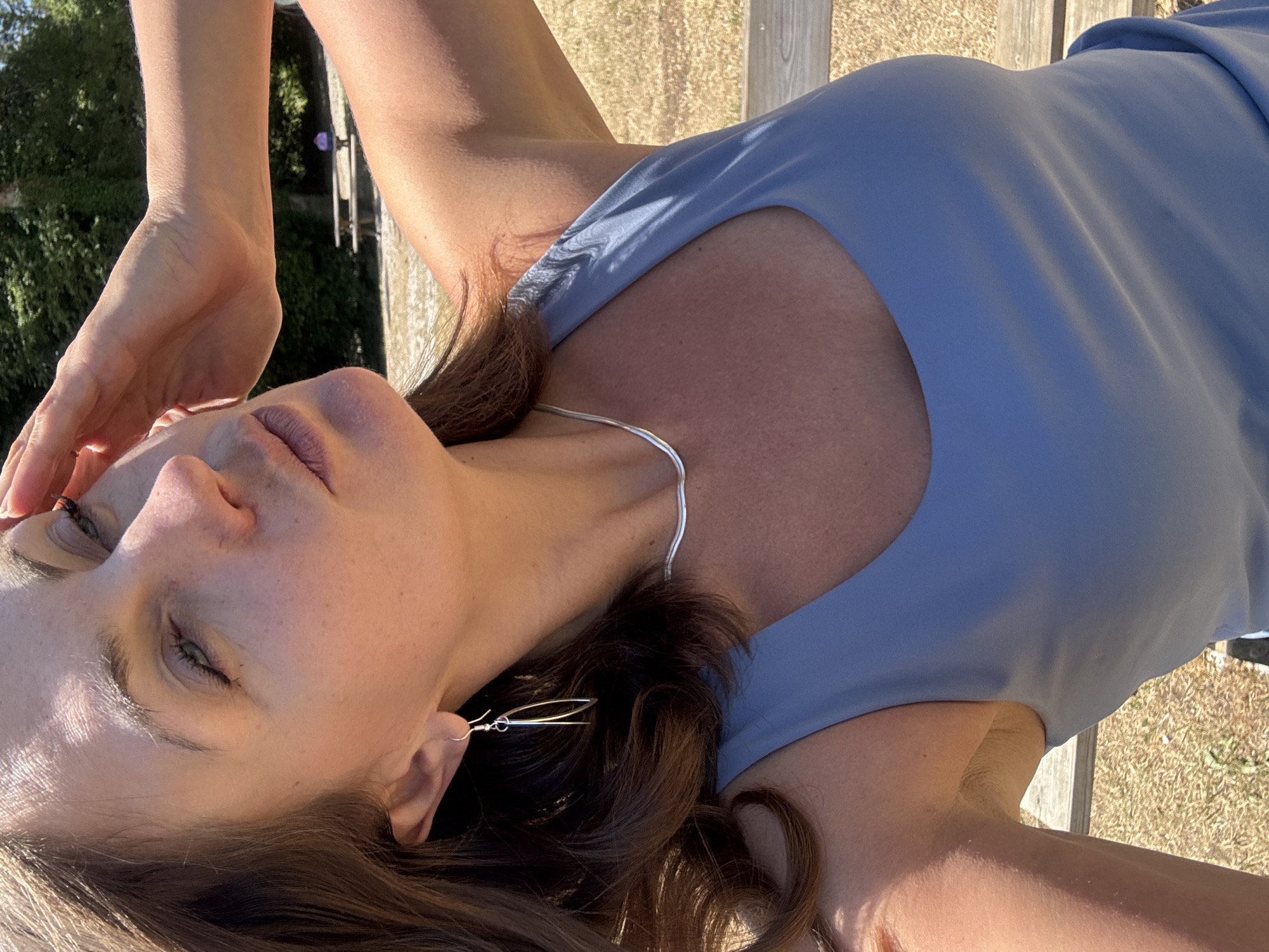 A woman is pictured from the chest up, outdoors on a sunny day, with her head tilted back and her right hand shielding her eyes from the sun. She has brown hair and is wearing a blue sleeveless top, a silver necklace, and silver earrings. The background shows a wooden bench and a dry, grassy area with trees and foliage. A distant picnic table with a person is visible. The setting appears to be a park or a natural area.