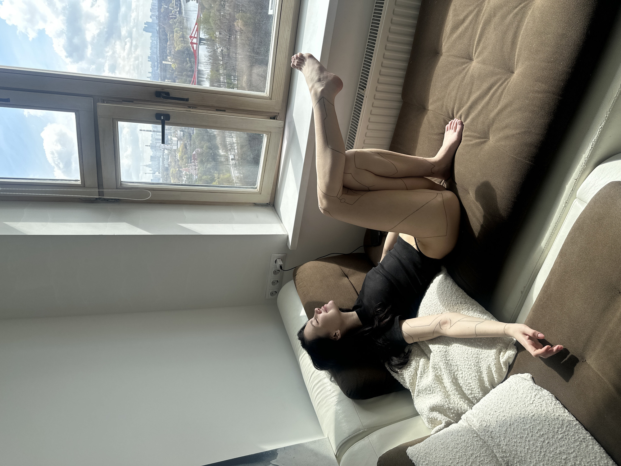 The image captures a woman relaxing on a sofa or daybed positioned alongside a large window, offering a panoramic view of a city. The woman is lying on her back, with her head resting on a pillow and her eyes closed, suggesting a moment of rest or contemplation. Her legs are extended towards the window sill, with one foot resting on it. Distinctive geometric line patterns are visibly drawn or tattooed on her arms and legs, adding a unique, stylized element to her appearance. She is wearing a dark, sleeveless top.
Bright natural light streams in through the window, casting strong shadows and highlighting the textures of the sofa and white, textured pillows. The sky outside is partly cloudy, and the city skyline of Khimki, Russia, is visible in the distance, featuring buildings, trees, and what appears to be a river with a red structure (possibly a bridge) spanning it. The window frame is made of light-colored wood, and one pane is slightly ajar. A white radiator is visible below the window. The sofa features a tufted brown section and a white armrest. A power outlet with multiple sockets is visible on the wall near her head.
The overall atmosphere is one of peaceful relaxation in a sunny indoor setting during the daytime. There is no visible text in the image.