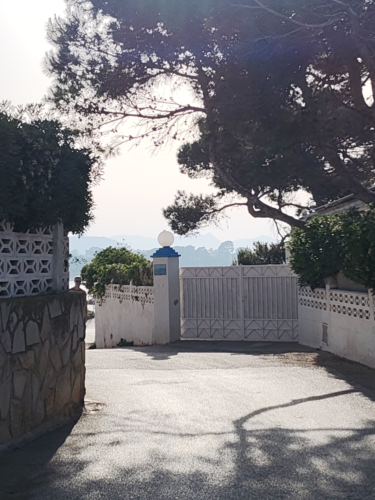 A view of a street and gated property in Rotes, Spain on a bright, sunny day. The foreground asphalt street is cracked and patched, with shadow patterns from trees above. A wall of quarried stone is visible on the left, adjacent to a section of wall made of patterned concrete blocks. In the center is a gated entrance to a property, flanked by white stucco walls topped with concrete blocks. A light fixture is visible atop a post at the gate. In the background, hazy mountains and sky are visible. A partially obscured person stands to the left of the gate entrance. A variety of trees and shrubs are scattered throughout the scene.