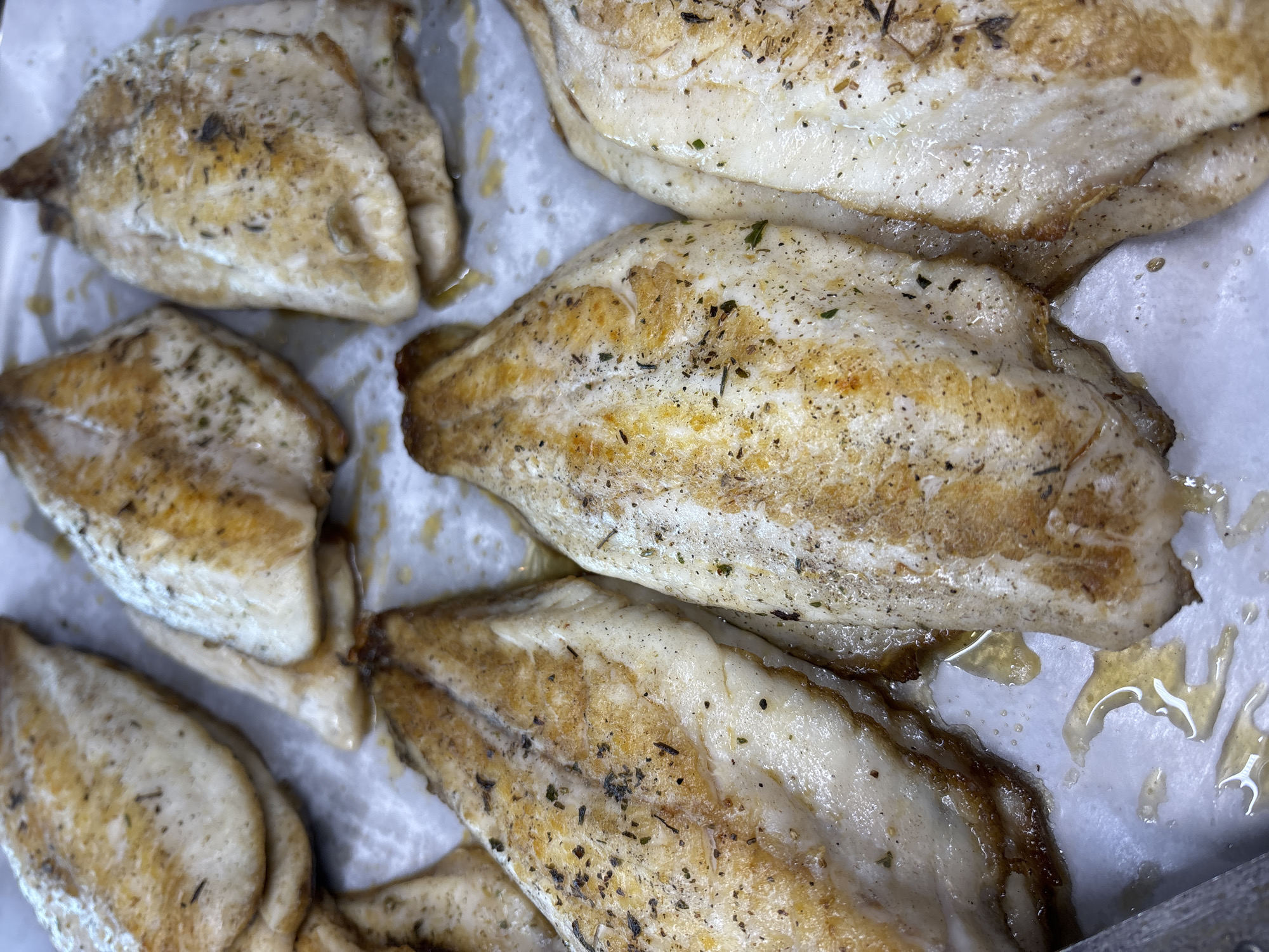 The image shows a close-up of cooked fish fillets arranged on white parchment paper in a tray. The fish appears seasoned with herbs and spices. There is visible oil or liquid in the tray with the fish. There is no indication of location or setting within Sofia, Bulgaria.