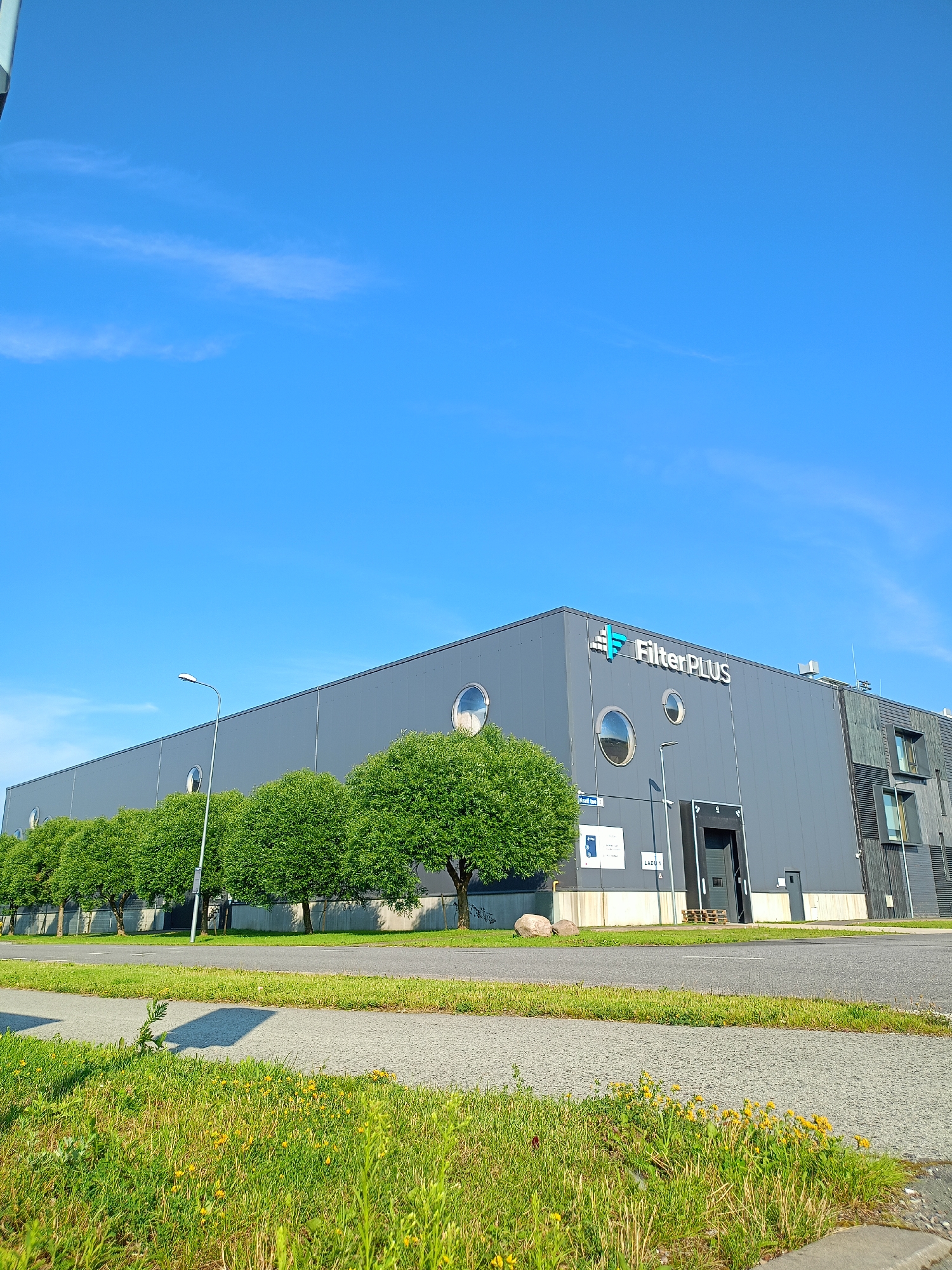 An exterior, low-angle, full shot shows a modern, grey industrial building with the name "FilterPLUS" in large white lettering on its facade. The building has a dark, slatted section on its right side, and a series of circular windows along its upper portion. A large, dark loading bay door is visible in the center of the building. In front of the building, a row of evenly spaced, rounded green trees lines the property. A streetlight stands in front of the trees. A paved road runs in front of the trees, and a strip of grass with small yellow wildflowers separates the road from another paved surface in the foreground. The sky is a clear, bright blue with a few wispy white clouds. The scene appears to be located in Peetri alevik, Estonia, on a sunny day.