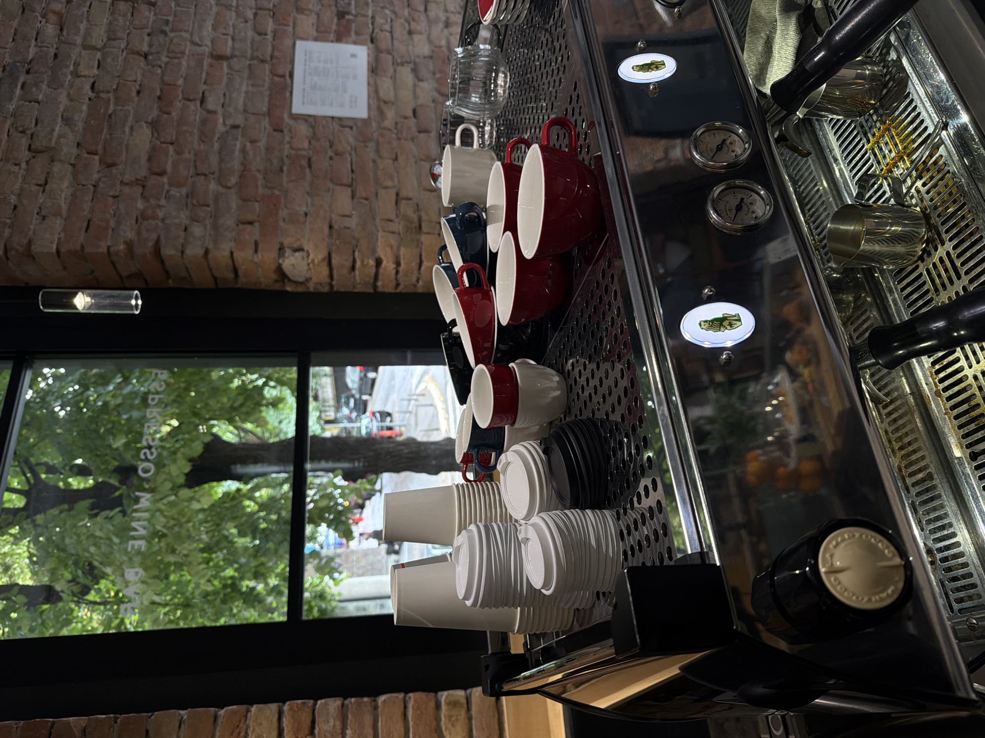 The image depicts the interior of a cafe, likely an espresso bar, with a prominent espresso machine in the foreground. The machine is made of stainless steel and features gauges, knobs, and various brewing accessories such as portafilters and milk pitchers. Stacked on the machine's surface are numerous paper cups and lids, as well as a collection of ceramic mugs in red, blue, and white. Behind the machine, a large window reveals an outdoor scene with trees and a street, suggesting an urban setting. The background is characterized by exposed brick walls and a modern pendant light fixture. A sign on the window, partially obscured by reflections, reads "ESPRESSO WINE BAR". The overall impression is one of a functional and stylish cafe environment, ready for service. The location of this cafe is in the city of Belgrade, Serbia.