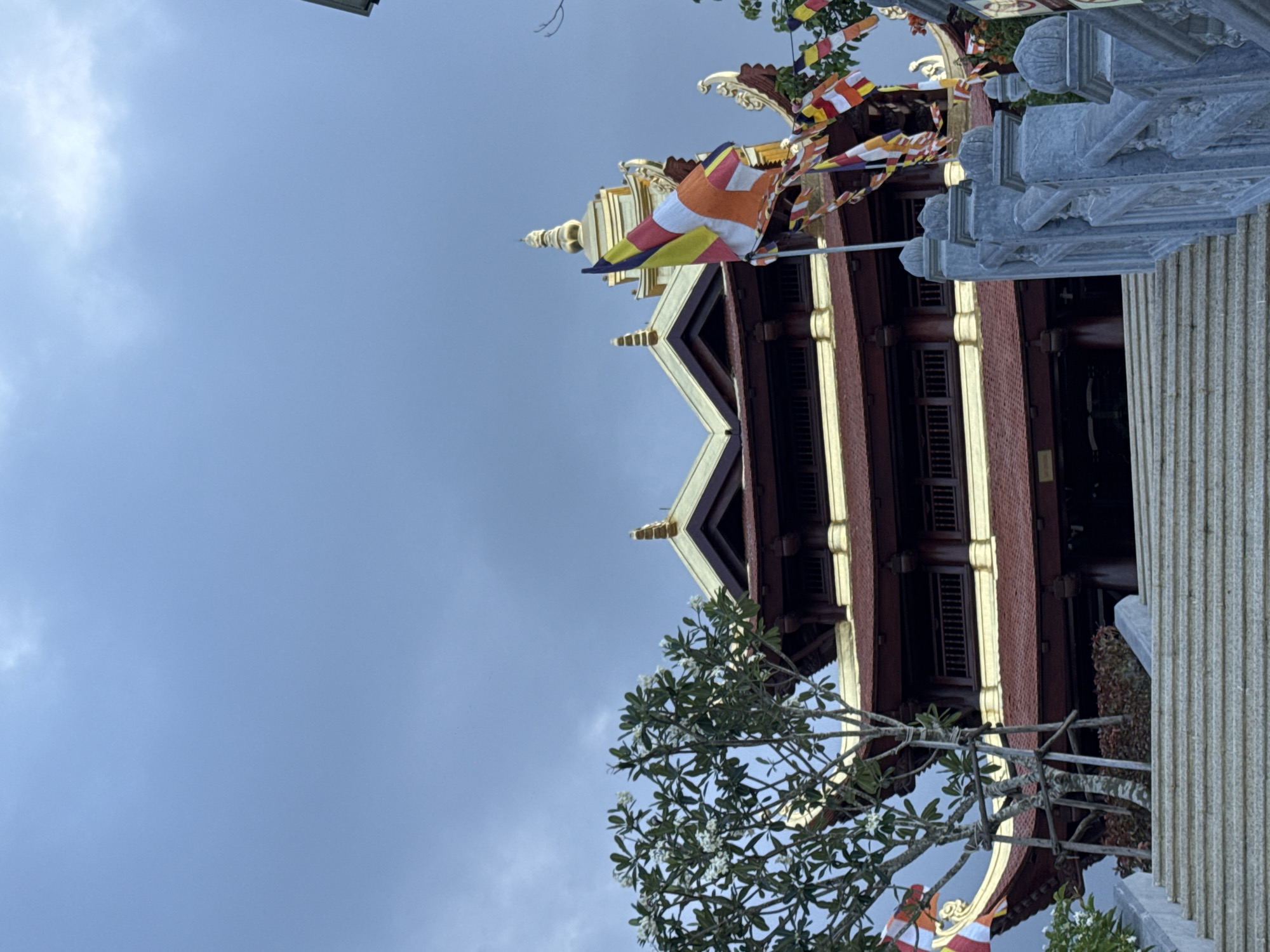 The image shows the exterior of a Buddhist temple, likely located in Diên Khánh, Vietnam. The temple features traditional architecture with tiered, dark brown wooden roofs trimmed in gold, and a prominent golden spire. Several colorful prayer flags are strung across the scene, fluttering in the wind. A stone staircase leads up to the temple entrance, flanked by ornate stone railings. To the left of the stairs, a flowering tree with white blossoms adds a touch of greenery. The sky is overcast with gray clouds, suggesting a muted or impending weather change. The scene is devoid of people, focusing entirely on the architectural and atmospheric elements of the location.