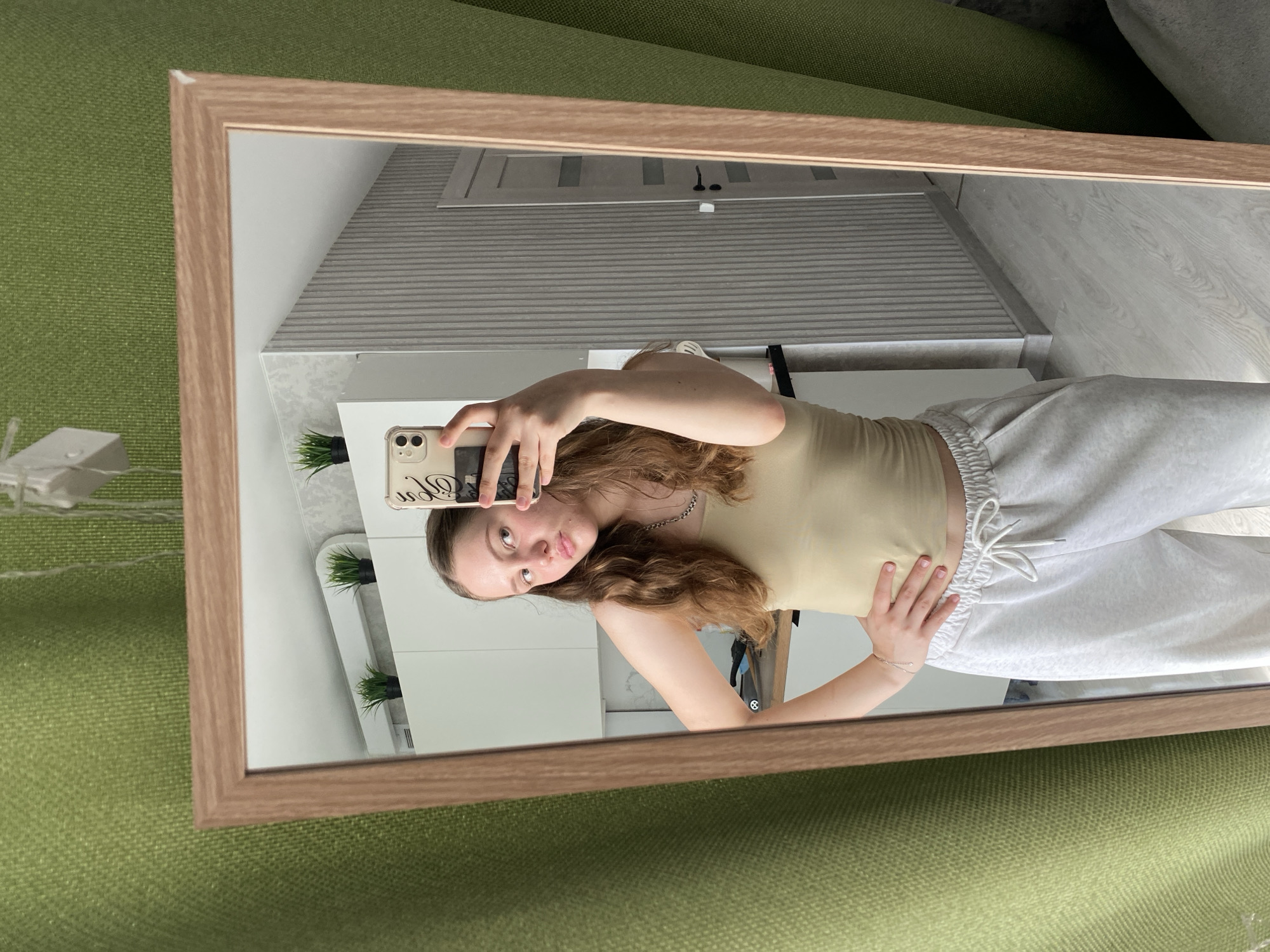 A young woman with long, wavy brown hair is taking a mirror selfie in what appears to be a modern kitchen or hallway. She is wearing a light beige tank top and light grey sweatpants with a drawstring. Her left hand is on her hip, and she is holding a smartphone in her right hand, angled to capture the reflection. The mirror is framed in a light wood. In the reflection, behind her, are white cabinets, a light gray wall with vertical ridged paneling, and a closed door. Small potted plants are visible on top of the cabinets. The overall environment suggests an indoor residential setting. There is no discernible information to indicate this location is in Izhevsk, Russia.