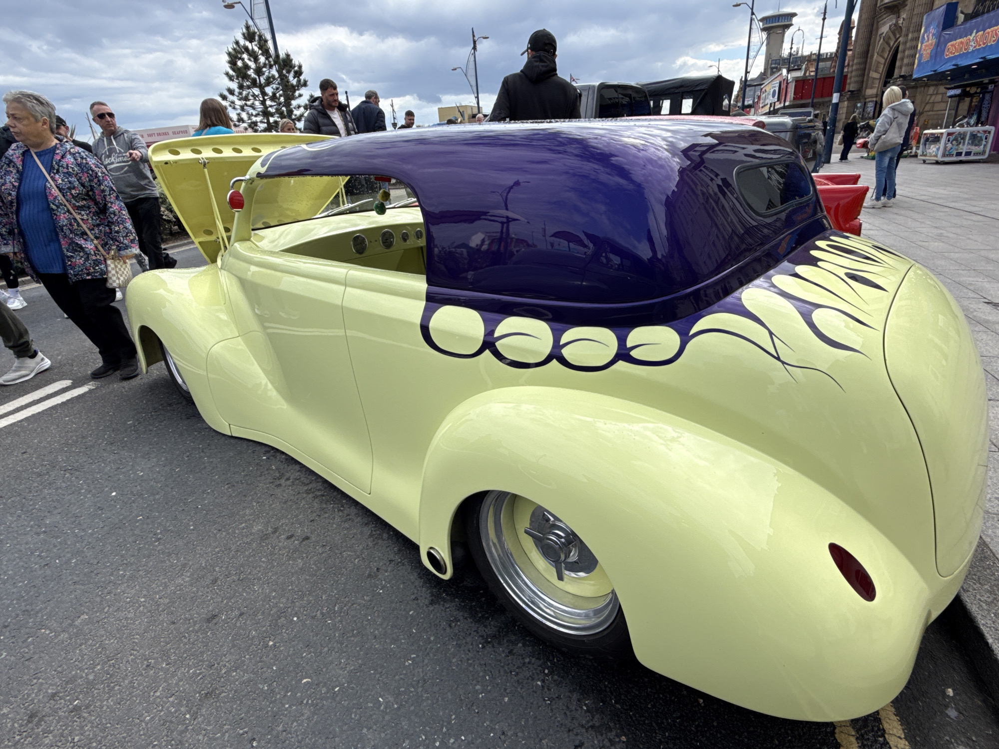 The image shows a custom-built, pale yellow hot rod with a dark purple roof and flame decals parked on a street in Great Yarmouth, United Kingdom. People are seen walking around and observing the car. The hood of the car is open. In the background, buildings, street lights, and signage suggest a seaside resort location. One sign reads "CASINO SLOTS." A woman with a floral jacket is standing close to the car on the left. Other onlookers stand or pass by on the pavement and road. The sky is overcast.