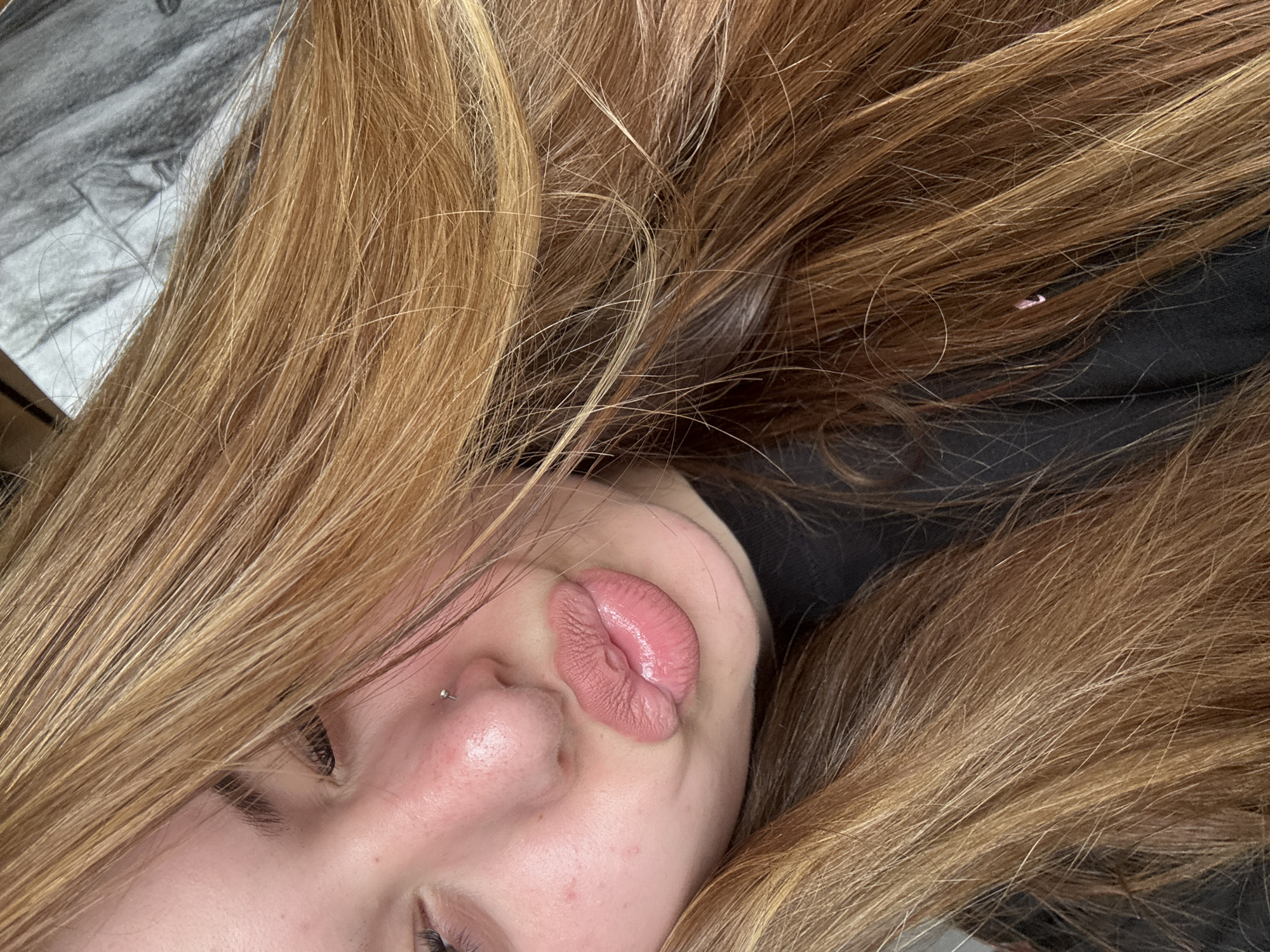A close-up selfie shows a person lying down with their head tilted. The individual has long, light brown hair partially obscuring their face. Visible facial features include closed eyes, a nose with a piercing, and pursed lips. The background is blurred and indistinct, showing hints of grey and black. There is no indication of Troyan, Bulgaria.