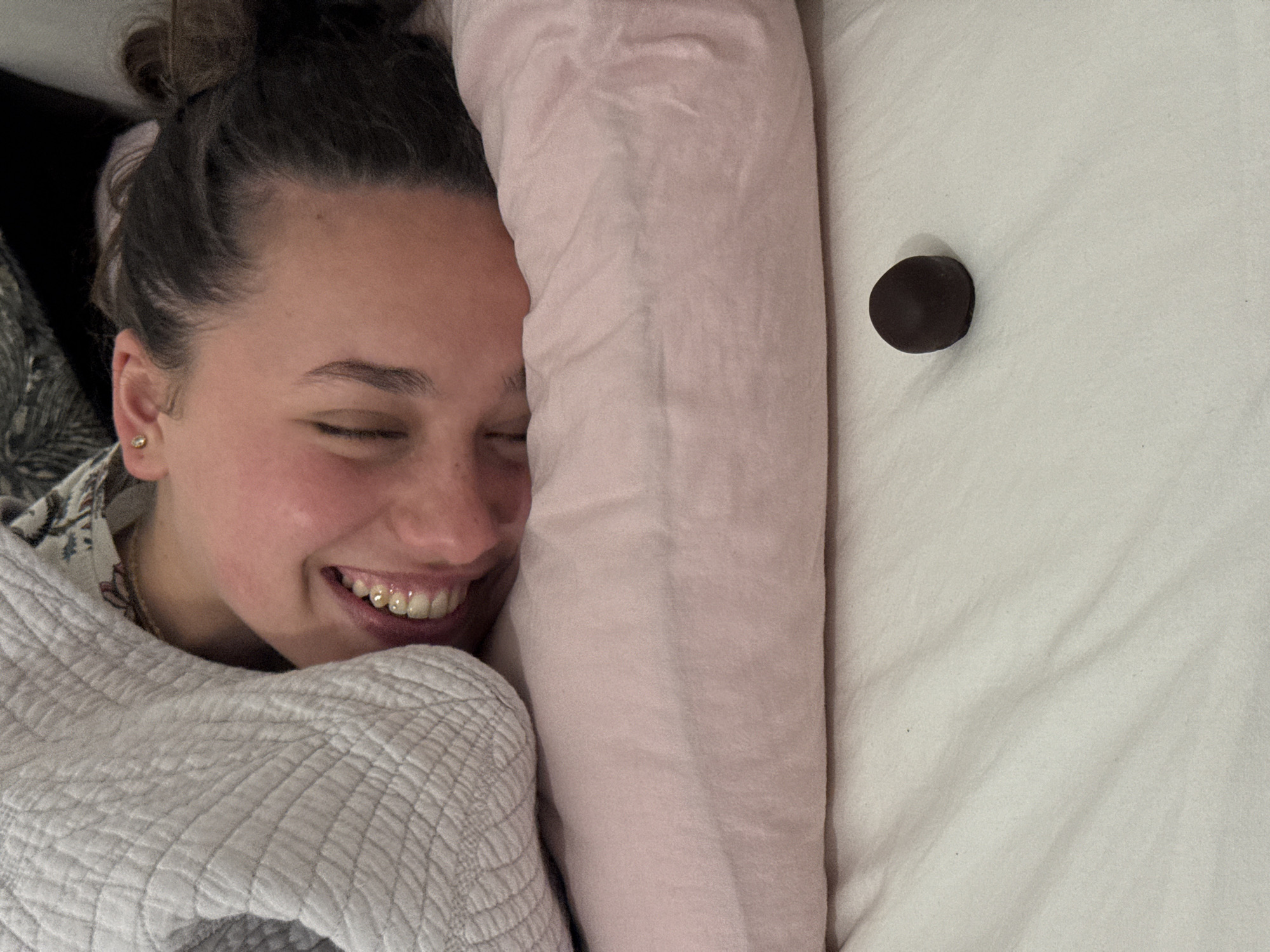 A person lies in a bed next to a pink pillow and a single, round, dark chocolate candy on a white sheet. The person is fair-skinned with brown hair in a messy bun and is wearing a white quilted blanket and a patterned shirt. The person's eyes are closed, and they are smiling, revealing gold dental work on their teeth. The location is not possible to determine from this image.