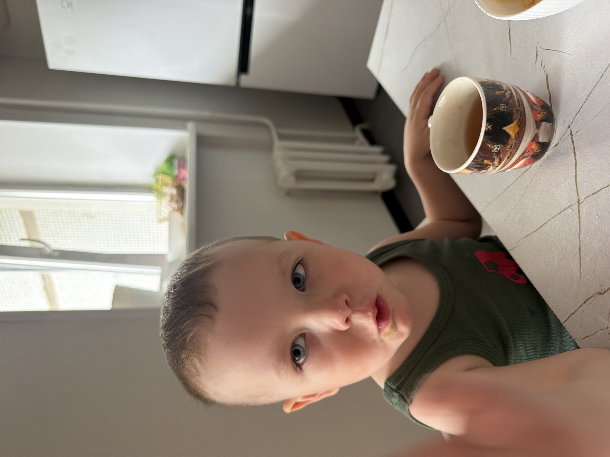 The image depicts a young child seated at a table indoors, likely within a residence in Chaikovsky, Russia. The child, with light skin, short brown hair, and blue eyes, is wearing a dark green ribbed tank top featuring a red stylized car graphic on the lower left front. Yellow food residue is visible on the child's chin and around the mouth. The child's left hand is extended into the foreground, partially obscuring the lower left corner of the frame, while the right forearm rests on the light-colored table. The child gazes directly at the camera with a slightly open mouth.
On the table, to the child's right, a white ceramic mug with a multi-colored panoramic illustration, possibly depicting a festive cityscape or temple scene, holds a light brown liquid consistent with tea. A portion of a white bowl is also visible on the far right edge of the table.
In the background, a window with a light-colored frame and translucent coverings allows natural light into the room. On the windowsill below the window, several small, clear glass bowls and other containers are present, holding various small items, possibly snacks or ingredients. Below the windowsill, a white, finned radiator is mounted on the wall. To the right of the radiator and window, a modern, two-door white refrigerator with dark handles is visible. The walls are light-colored, and the floor appears to be a dark grey.