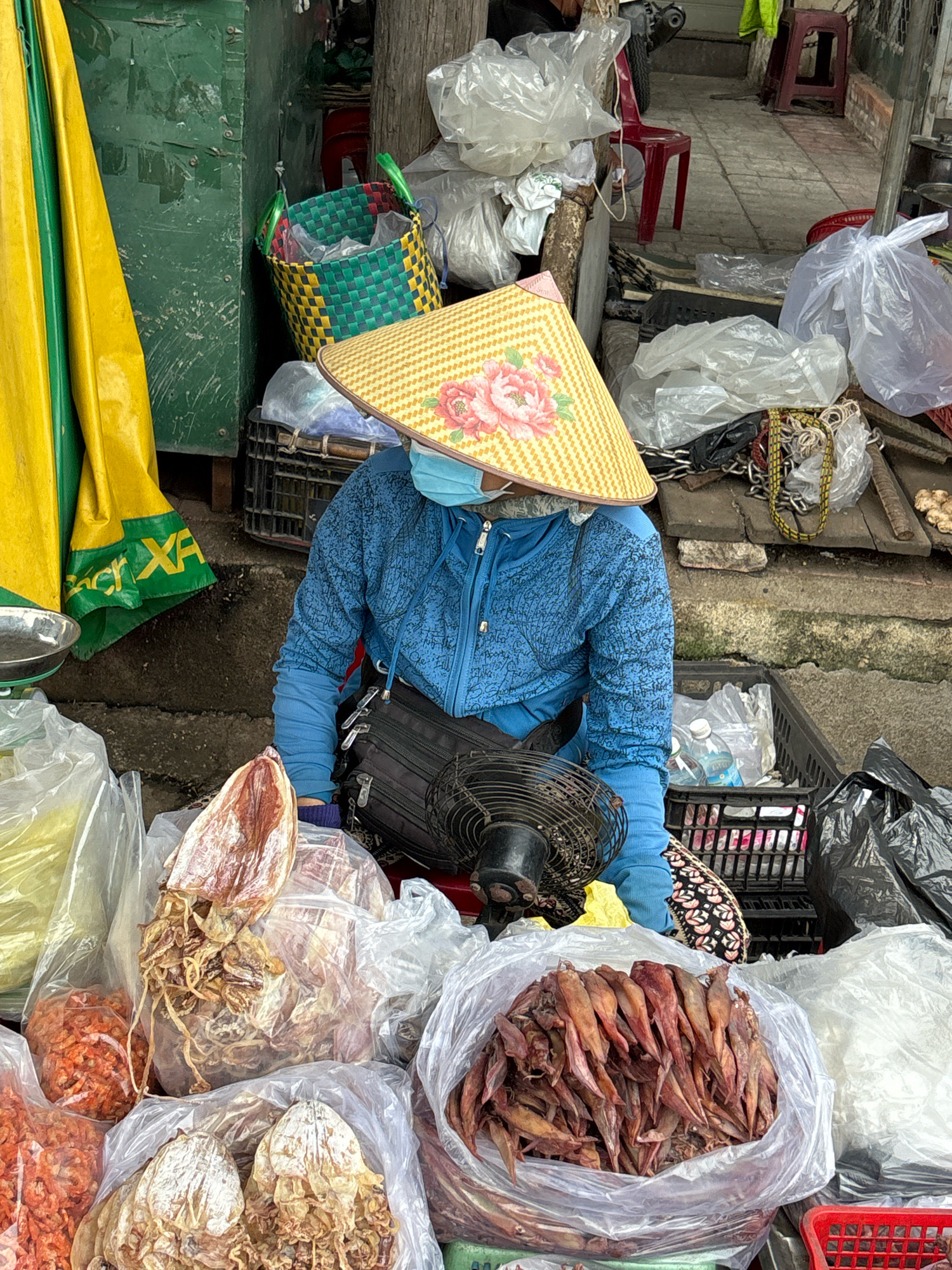 This is a street market scene in Nha Trang, Vietnam. A person wearing a blue jacket, a light blue mask, and a conical straw hat with a floral pattern is seated behind a stall. The stall displays various dried seafood items, including large dried squid and small dried shrimp, packaged in plastic bags. A black electric fan is positioned in front of the seated person. In the background, there are green and yellow tarps, a green metal cabinet, plastic bags, and stacked crates. The ground appears to be a paved walkway.