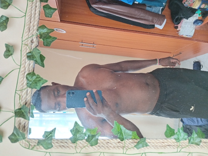 A man takes a selfie in a mirror. He is shirtless, wearing black athletic shorts. The mirror has a rope-like frame decorated with artificial ivy. Behind him is a wooden cabinet or dresser, with clothes piled on top. A water bottle or mug is also visible. The man is holding a blue smartphone.
