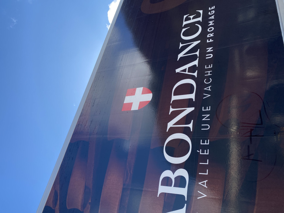 The image depicts a large outdoor banner or sign, partially visible against a bright blue sky with sparse white clouds. The banner features dark brown background imagery, resembling stacked wooden planks or blocks. Prominently displayed in white capital letters is the word "ABONDANCE". Below it, in smaller white capital letters, is the French slogan "UNE VALLÉE UNE VACHE UN FROMAGE" (A Valley A Cow A Cheese). To the right of "ABONDANCE", a red shield logo with a white cross is visible. Faint, dark graffiti-like markings are present on the lower left section of the banner, partially obscuring the background. The photograph is taken from a low angle, looking upwards at the sign and the sky. This banner is located in the city of Abondance, France, promoting the local region and its famous cheese.
