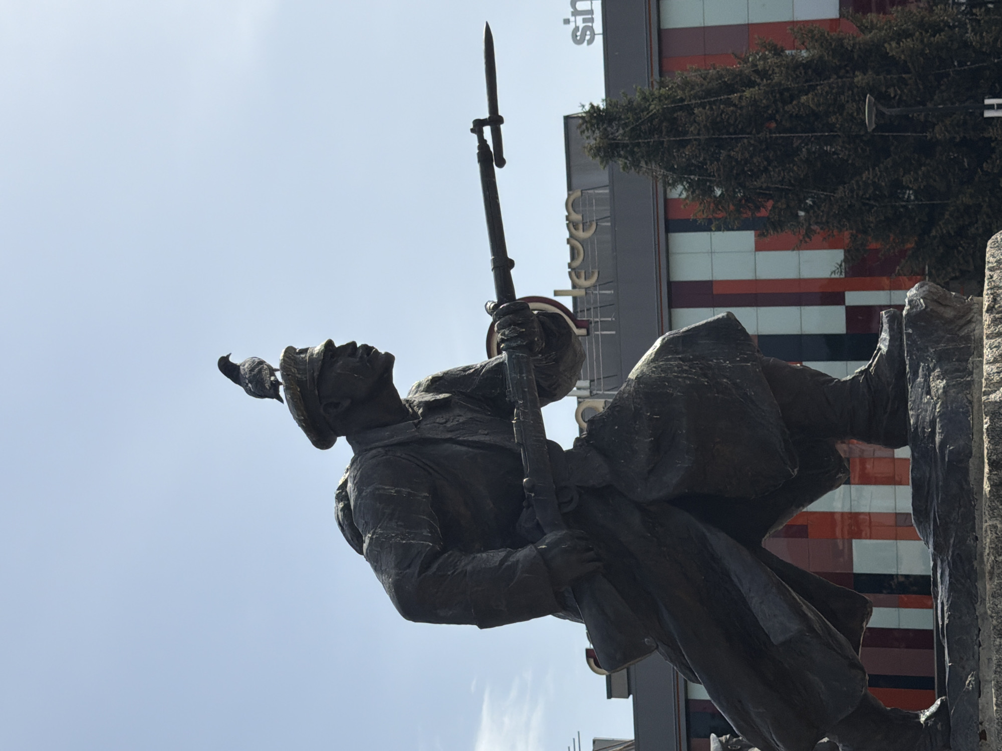 The image shows a statue of a soldier holding a rifle with a bayonet attached. A bird is perched on the soldier's hat. The statue is situated outdoors, with a building in the background displaying a pattern of red, white, and gray rectangles. The word "Leeses" can be seen vertically on the building in large letters. The photo is likely taken in Pleven, Bulgaria, where a monument depicting a soldier with a rifle exists.