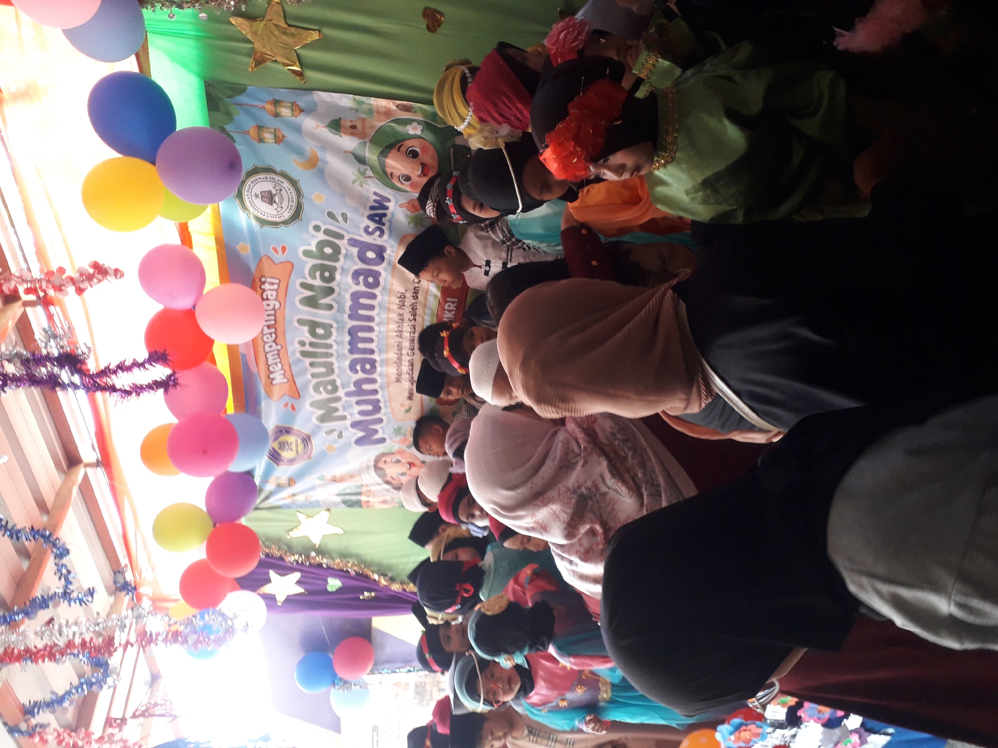 A group of children, dressed in traditional Islamic attire, are gathered in a room decorated for a Maulid Nabi celebration. The room features balloons, tinsel, and a banner that reads "Memperingati Maulid Nabi Muhammad SAW" (Commemorating the Birthday of Prophet Muhammad SAW). The children appear to be participating in or observing an event, possibly a religious gathering or school function, held in Cankuang, Indonesia.