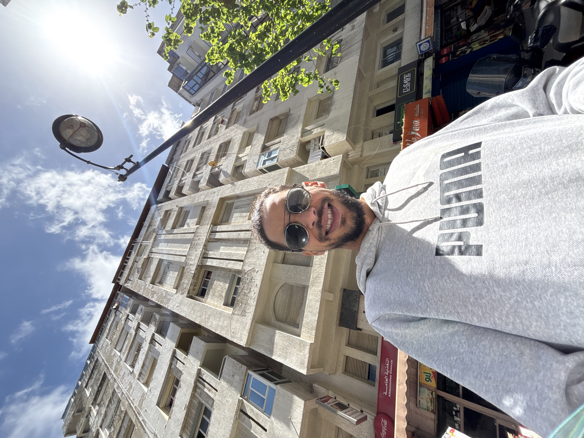 The image presents a low-angle selfie of a smiling adult male in Tanger, Morocco, taken on a bright, sunny day. The man, with short dark hair, stubble, and reflective aviator sunglasses, wears a light grey hooded sweatshirt emblazoned with "PUMA" in black capital letters. He is positioned in the foreground, looking directly at the camera.
Behind the man, a tall, multi-story building with a weathered, light-colored facade rises vertically, dominating the background against a blue sky with scattered white clouds. The building features numerous windows, some with blue glass, and balconies. At its base, visible street-level storefronts include a red Coca-Cola sign, a red awning with "التغذية العامة" (Alimentation Générale - General Food/Store) in Arabic, and a green-and-orange sign partially displaying "U-SAVE." A black street lamp with a circular luminaire extends into the frame from the right, and a tree with vibrant green foliage is visible on the far right. The sun is prominent in the upper right, casting a strong glare. Part of a dark-colored motorcycle or scooter is visible on the far right, near the bottom.