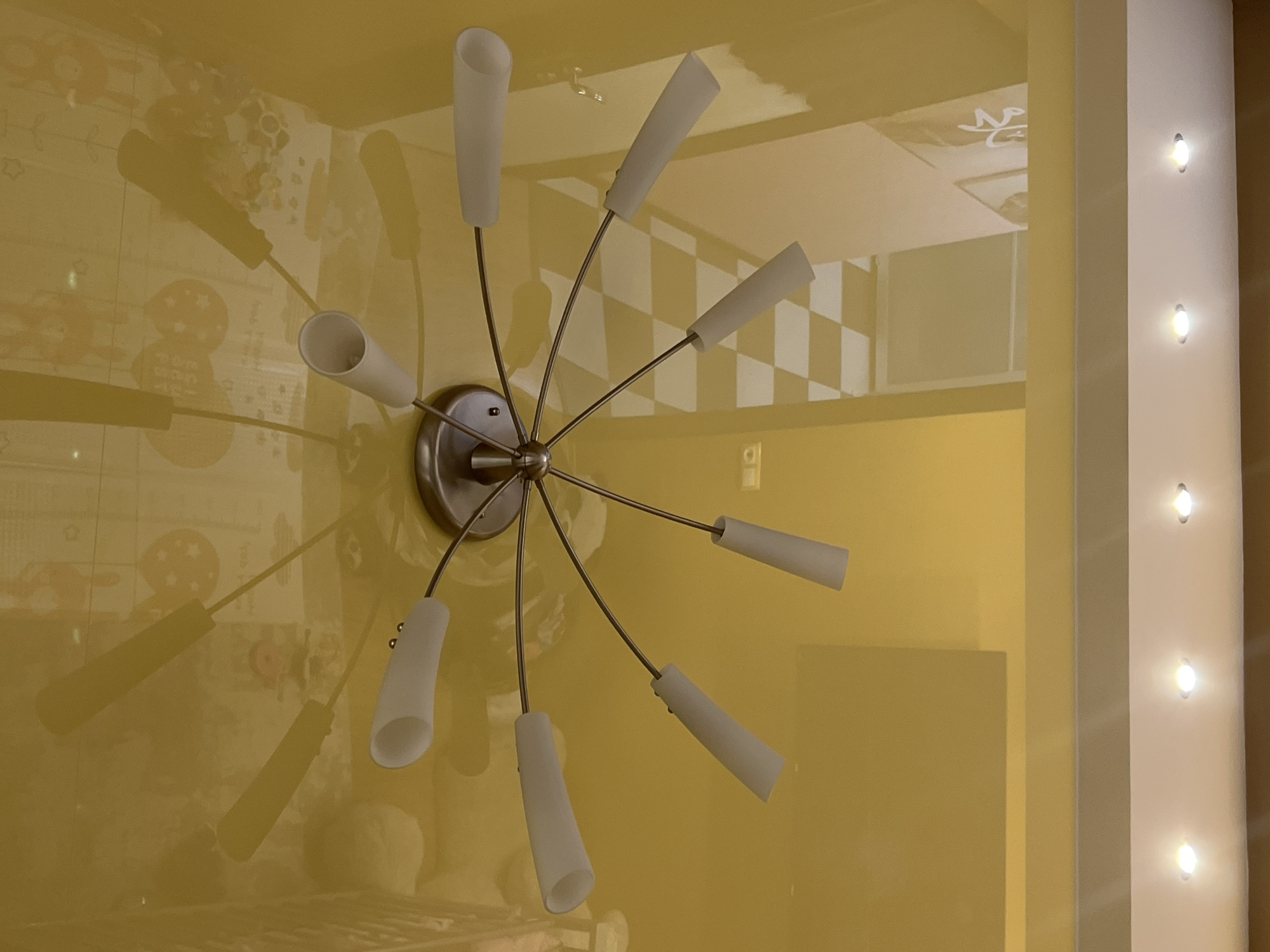 The image captures an indoor scene, primarily featuring a modern ceiling light fixture against a glossy, reflective yellow ceiling. The light fixture has a brushed metal base from which several thin, curved arms extend, each holding a white, conical light shade. Eight shades are clearly visible, housing illuminated bulbs.
The highly reflective yellow ceiling acts as a mirror, showing distorted reflections of the surrounding room. To the left of the main fixture, reflections reveal patterns and shapes, including stars and what appear to be outlines of figures or objects often found in a child's room. More broadly, indistinct reflections of other room elements are scattered across the left side of the ceiling.
To the right, a significant portion of the reflection shows a white wall with a distinctive checkerboard pattern. Adjacent to this, a vertical white panel equipped with five evenly spaced, recessed spotlights casts bright, warm light into the space, providing additional illumination. Above the checkerboard pattern in the reflection, a faint, stylized symbol or text, possibly resembling Arabic script, is discernible on a white surface. Other extremely faint and illegible text, potentially including English words like "ON ME" or "IN ME" and some numbers, can be vaguely seen in other reflections.
The room is artificially lit, suggesting it is evening or night. The overall impression is of a well-lit interior space. This scene is situated in Yekaterinburg, Russia.