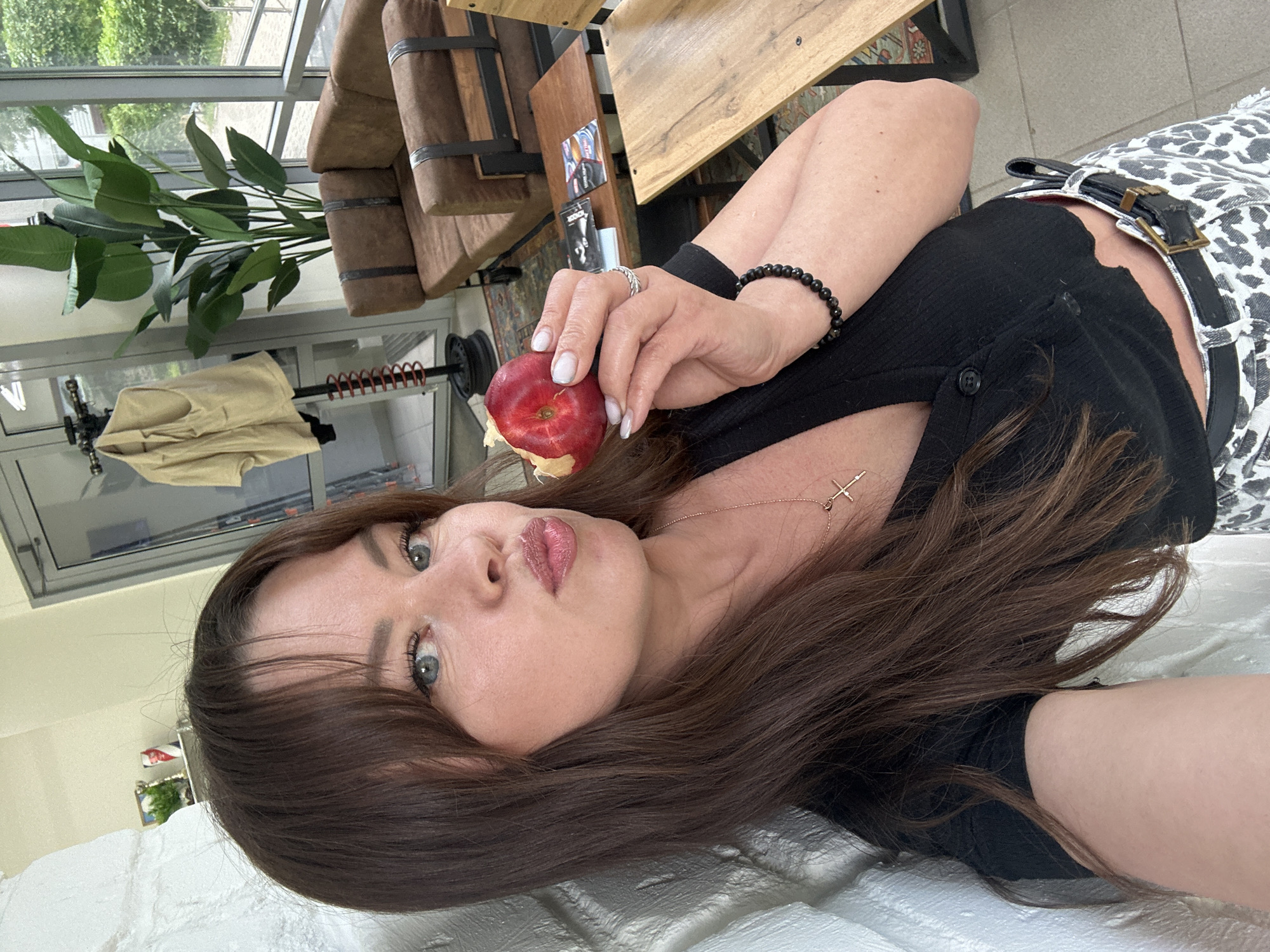 A woman is holding a nectarine to her face, her lips pursed. She has long brown hair and is wearing a black crop top, a silver necklace with a cross, and a patterned skirt with a black belt. She is standing in front of a white brick wall. In the background, there is a large plant, a brown sofa, and a glass door that reveals an outdoor area with greenery. The scene appears to be an indoor waiting area or lounge, potentially in a business or public space. There is no visible indication of the city of Chaikovsky, Russia.