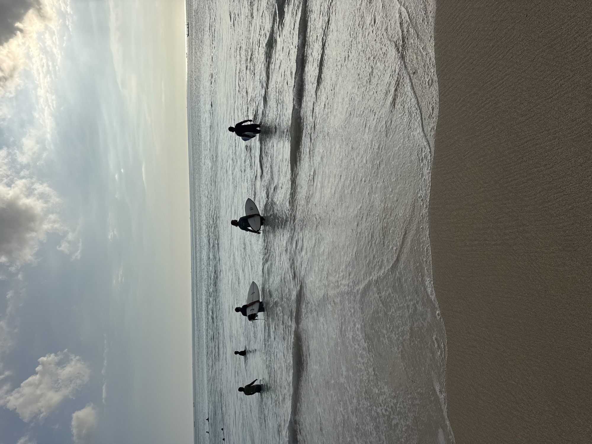 The image captures a beach scene with several individuals in the water, likely surfers preparing to ride waves. The beach is located in Kuta Selatan, Indonesia, as indicated by the sandy shoreline and the ocean. Several surfers, are scattered in the water, some with surfboards. The sky overhead is partly cloudy, providing a soft and diffused light. The water surface shows gentle undulations. The beach sand appears dark and wet near the water's edge.