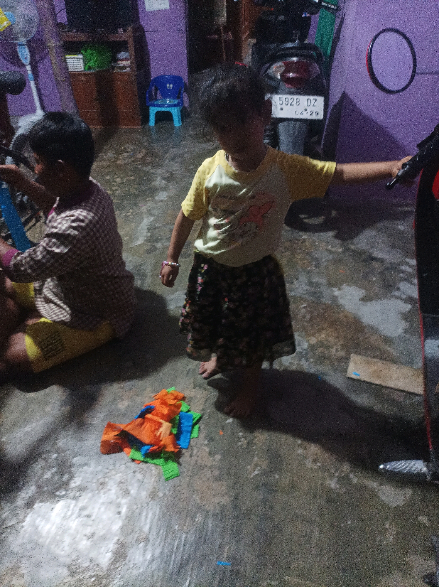 In the city of Canga'an, Indonesia, two children are present in what appears to be a living room or garage area. The child on the left is seated, wearing a checkered long-sleeved shirt over a yellow garment and possibly yellow shorts. This child is looking to the left and appears to be interacting with something out of frame. The child on the right is standing, wearing a yellow t-shirt with graphics and a dark, floral-patterned skirt. This child is facing forward, with their right arm extended towards the viewer. In front of the standing child, a pile of brightly colored paper, likely from a craft project, is scattered on the floor. To the right of the standing child, a portion of a motorcycle is visible. The room itself has a dark, textured floor. In the background, a blue plastic chair is visible, along with furniture and a standing fan. The walls are painted in shades of purple and have various objects mounted on them, including what looks like a circular mirror.