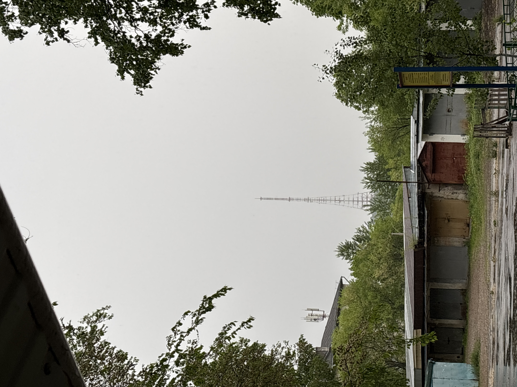 The image depicts an outdoor scene in an urban environment. The sky is overcast and a tall lattice tower is visible in the distance, rising above a line of trees. In the foreground, there are several low buildings, possibly garages or storage units, arranged in a row. A sign or informational board is mounted on a blue frame next to one of the buildings. Trees frame the upper portion of the image, and vegetation is present alongside the buildings. The ground appears damp, suggesting recent rain. A fragment of a building roof is visible in the lower left corner. The location is Unknown, Unknown.