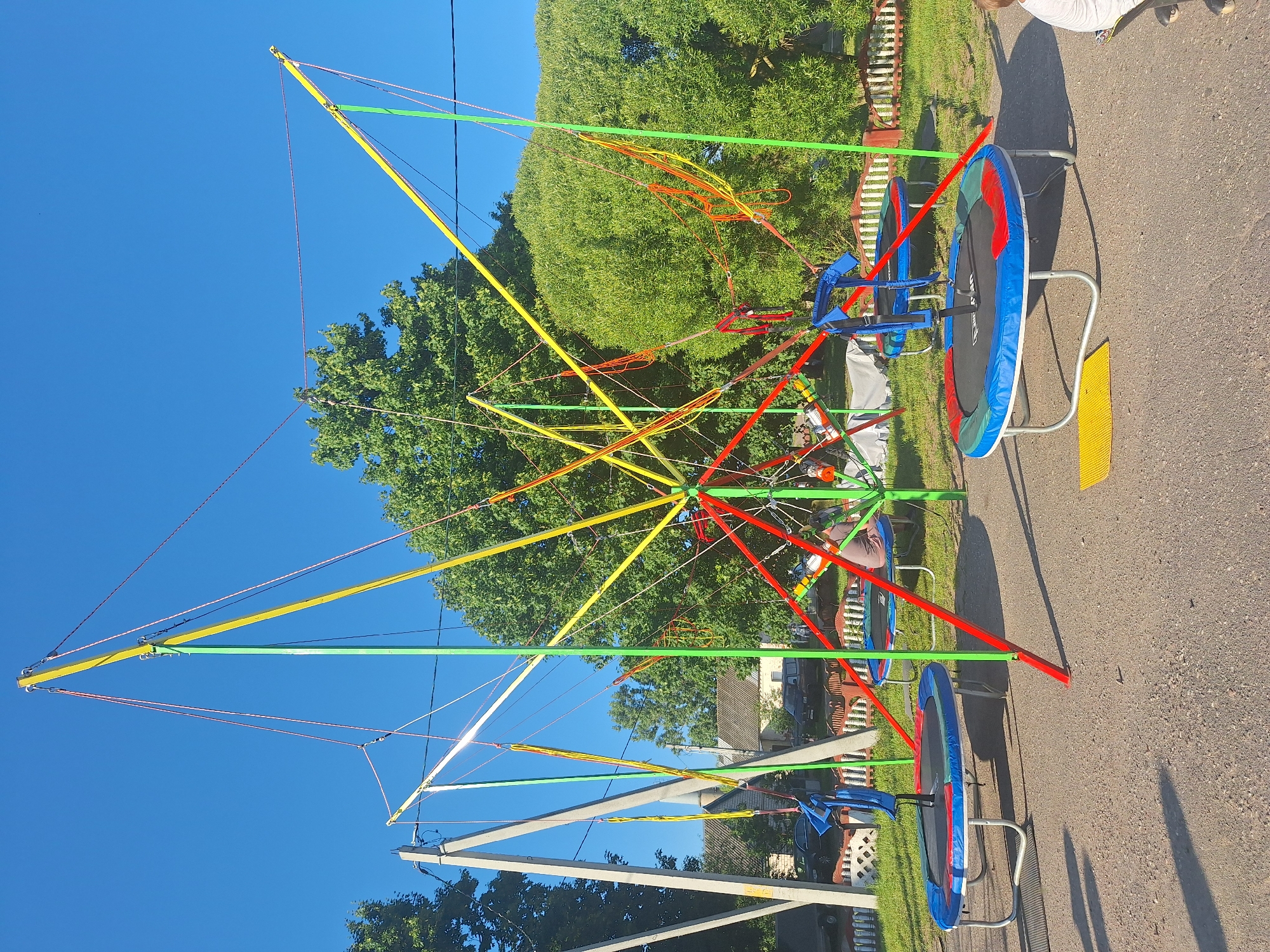 An outdoor scene captured under a clear blue sky features a bungee trampoline setup. The main structure is a colorful framework of red, yellow, and green poles and ropes, from which several bungee cords are suspended. Two trampolines are positioned on the ground beneath the bungee apparatus. The trampoline in the foreground is blue with red and green accents, equipped with a blue harness. Another trampoline, similar in design, is visible further back. In the background, there are trees, houses, cars, and a white picket fence. A person can be seen sitting on the rear trampoline, and a portion of another person is visible on the right side of the frame, facing away from the camera. The ground is covered with gravel and asphalt. The location is identified as Цімкавіцкі сельскі Савет, Belarus.