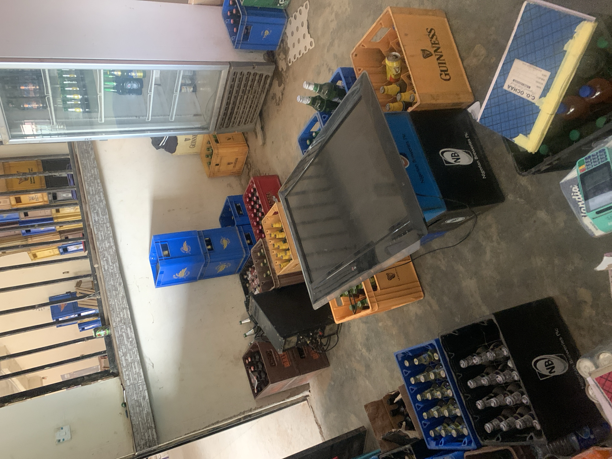 This image depicts an indoor storage or retail area for beverages in New Stadium, Nigeria. The concrete floor appears worn and soiled in areas. Walls are painted a light color, with a tiled section on a raised ledge or counter visible in the upper left quadrant. Metal bars are installed, likely part of a window or partition, behind which additional stacked crates are visible.
No individuals are present.
The scene is dominated by numerous crates of bottled and canned beverages. Blue plastic crates, many bearing a bird emblem and some from Nigerian Breweries Plc (NB), are stacked and dispersed throughout. Yellow/orange plastic crates, several labeled "GUINNESS," contain various bottles and a yellow beverage can. Brown wooden crates, some marked "MALTA," are stacked on the left, containing dark-bottled beverages. A red plastic crate, partially visible, appears to contain "AMSTEL" branded bottles.
A tall, single-door glass refrigerator stands against the right wall, displaying multiple shelves stocked with green-bottled beverages (likely Heineken) and darker-bottled beverages. A white, perforated plastic sheet, possibly beverage packaging, lies on the floor near the refrigerator.
In the central foreground, a black flat-screen display, likely a television, rests horizontally on a blue plastic crate, which in turn sits on a black crate branded "NB Nigerian Breweries Plc." To the left of this display, a black electronic unit, possibly an audio amplifier, is visible with attached wires.
Other notable objects include:
- A light switch on the upper left wall.
- A green can and a small silver object on the tiled ledge.
- A light-colored device labeled "Flandia," possibly a point-of-sale terminal, in the bottom left.
- A blue notebook labeled "C.O. OCHAA" placed inside a crate in the bottom right, alongside brown bottles.
- Empty cardboard boxes are also visible on the floor.
The overall environment suggests a commercial space dealing with beverage distribution or sales, possibly a bar or a wholesale outlet.