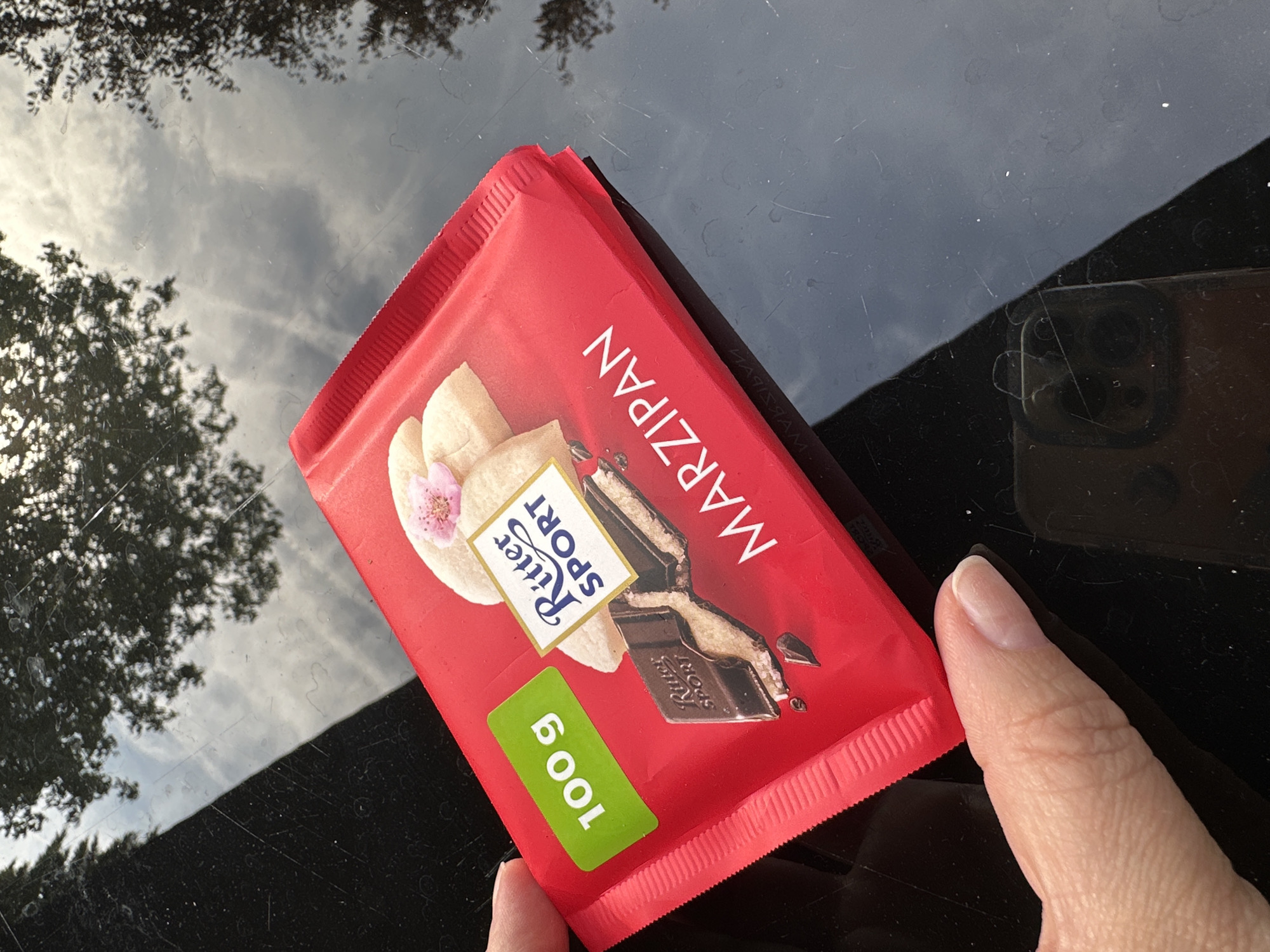 A close-up shot shows a person's thumb and index finger holding a red Ritter Sport Marzipan chocolate bar. The bar is square, as is characteristic of the brand, and features an image of the marzipan filling and chocolate. Text on the wrapper indicates "100 g" in a green sticker and "Ritter SPORT" above an illustration of the chocolate bar. The marzipan flavor is clearly stated as "MARZIPAN". The chocolate bar is placed on a dark, reflective surface, likely a table or countertop, which shows reflections of trees and the sky, suggesting an outdoor or semi-outdoor setting. There are some smudges and scratches visible on the reflective surface. The image does not provide specific information about a location in Krefeld, Germany.