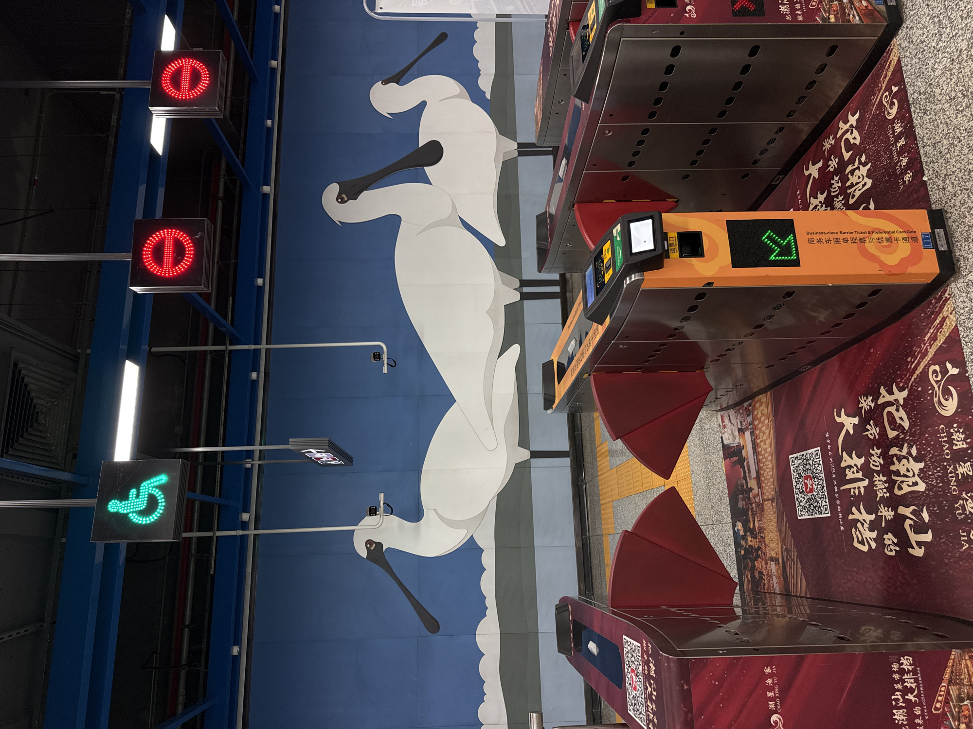 The image shows an interior view of what appears to be a subway station in Nanshan District, China. In the foreground, there are several ticket gates, some of which are open and illuminated with green lights, indicating they are operational. The ticket gates are adorned with red panels and advertisements featuring Chinese text and QR codes. Above the ticket gates, a mural adorns the blue wall, depicting three large, stylized white birds with long beaks, resembling spoonbills. Hanging from the ceiling are illuminated signs. One sign displays a green wheelchair symbol, indicating an accessible route. Two other square signs display red "forbidden" symbols. The overall environment is clean and well-lit, suggesting a public transportation hub.