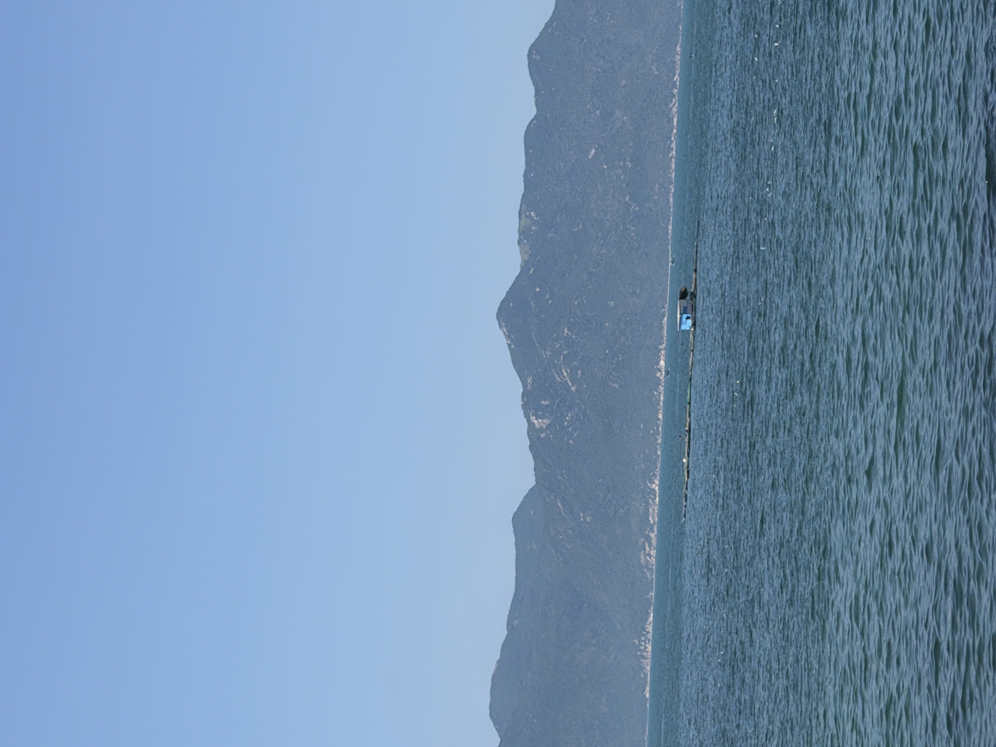 The image displays a maritime scene in Vĩnh Lương, Vietnam, under a clear, light blue sky. A continuous range of dark, heavily vegetated mountains forms the distant background, their lower slopes meeting a discernible rocky coastline. The expansive foreground and mid-ground are dominated by deep blue-green water exhibiting a rippled surface texture. In the mid-ground, a man-made floating structure is visible. This structure consists of a small, light blue, shed-like building with a dark roof, situated upon a dark, elongated platform or a series of interconnected rafts. This entire assembly extends horizontally across the water, consistent with marine aquaculture infrastructure. No individuals are visible within the frame. Scattered small white specks are present on the water's surface.