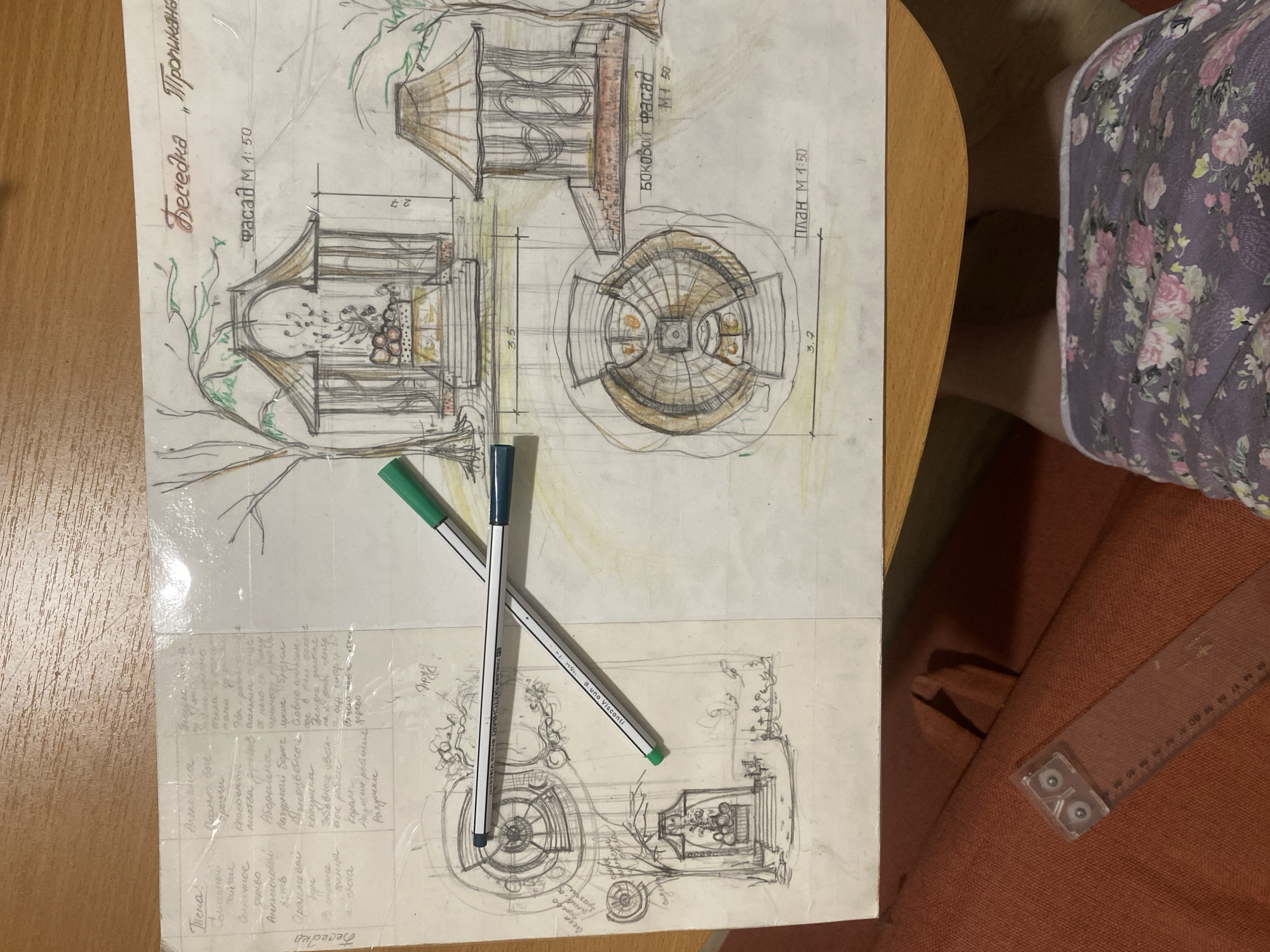 The image shows a hand-drawn architectural sketch on a white sheet of paper resting on a wooden table. The sketch depicts a gazebo design with the title "Gazebo 'Tropicana'" written in Russian. There are three views of the gazebo: a front facade, a side facade, and a plan view. The drawing includes dimensions and is labeled with scales (M 1:50). Two pens, one green and one blue, lie diagonally across the paper. Handwritten notes in Russian are visible on the lower left corner of the sheet. The table is partially visible, and in the background, there is a person wearing floral-patterned clothing and an orange chair. A clear plastic ruler with a case is also visible. The location is not discernible from the image, and no mention is made of Yekaterinburg, Russia.