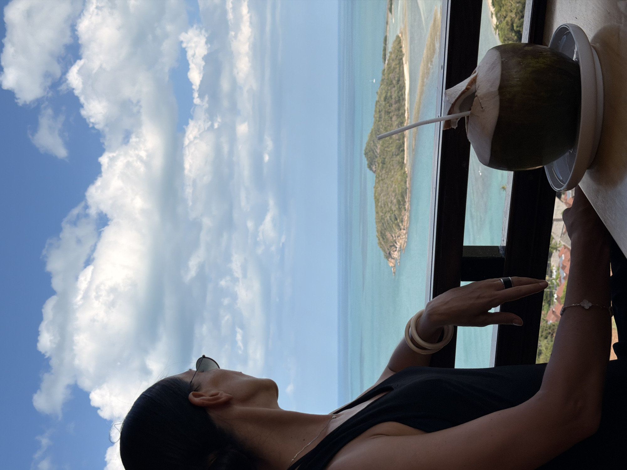 A woman with dark hair pulled back and wearing sunglasses gazes out at a tropical island and the turquoise ocean from a balcony in the Ko Samui District of Thailand. She is wearing a black tank top and several pieces of jewelry, including bangles on her wrist and a bracelet on her arm. In the foreground, a fresh coconut with a straw sits on a saucer on a wooden table. The sky is a brilliant blue, dotted with fluffy white clouds. The background shows a lush green island with a sandy spit connecting it to the mainland, and the clear blue water extends to the horizon.