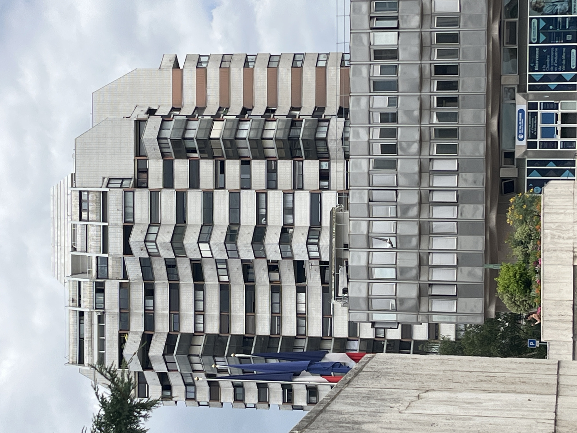 This is a frontal, eye-level shot of a multi-story building in Créteil, France. The building is composed of two distinct sections. The upper section is a tall, white, brutalist-style tower with a stepped facade featuring numerous balconies and windows. It has a white, corrugated material on the upper floors, interspersed with rows of windows. Some balconies have decorative elements. The lower section is a wider, gray, glass-fronted building with rows of rectangular windows, many of which are open. French flags are visible on the left side of the image, in front of the main building. In the foreground, a concrete wall occupies the left portion of the frame. Behind the lower building, there is greenery, including trees and flowers, and a person is visible standing near a parking sign. The sky is overcast. The signage on the lower building indicates it is the "Chambre de Commerce et d'Industrie du Val-de-Marne."
