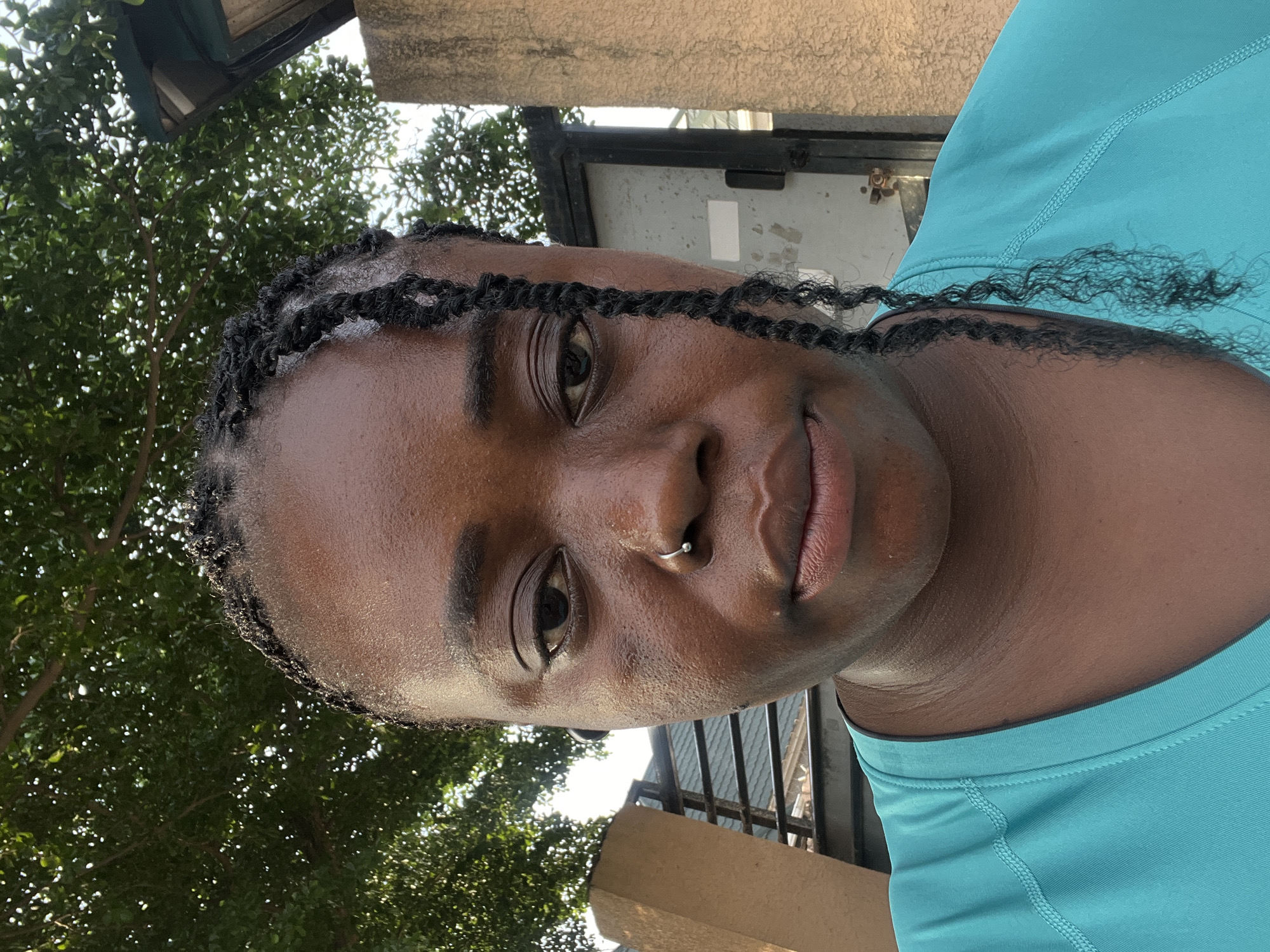 A close-up, frontal, outdoor selfie captures the upper body of a Black woman with braided hair and a nose ring. She is wearing a turquoise t-shirt. The background features lush green foliage from a tree, a textured concrete wall, and a dark metal gate with horizontal bars. The setting is consistent with a residential area in Abuja, Nigeria, likely during daylight hours given the natural light illuminating the scene.