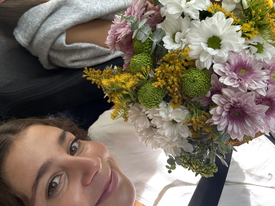 An image captures a close-up perspective inside what appears to be a vehicle. A female subject occupies the left side of the frame, partially visible from the chest upwards. She has medium brown hair, brown eyes, and is smiling towards the camera. She wears a white collared shirt, and a dark seatbelt crosses her chest from her right shoulder to her left hip. To her right, a second individual is partially visible, wearing a light gray hooded sweatshirt. In the foreground and central to the image is a vibrant bouquet of flowers. The bouquet contains white chrysanthemum-like flowers, pink chrysanthemums, bright yellow flowers (possibly solidago), and distinctive round, green-budded flowers (possibly green button chrysanthemums), interspersed with green foliage including eucalyptus leaves. The background is dark and blurred, consistent with the interior of a vehicle. No specific landmarks are visible to confirm the stated location of Aldoar, Foz do Douro e Nevogilde, Portugal.