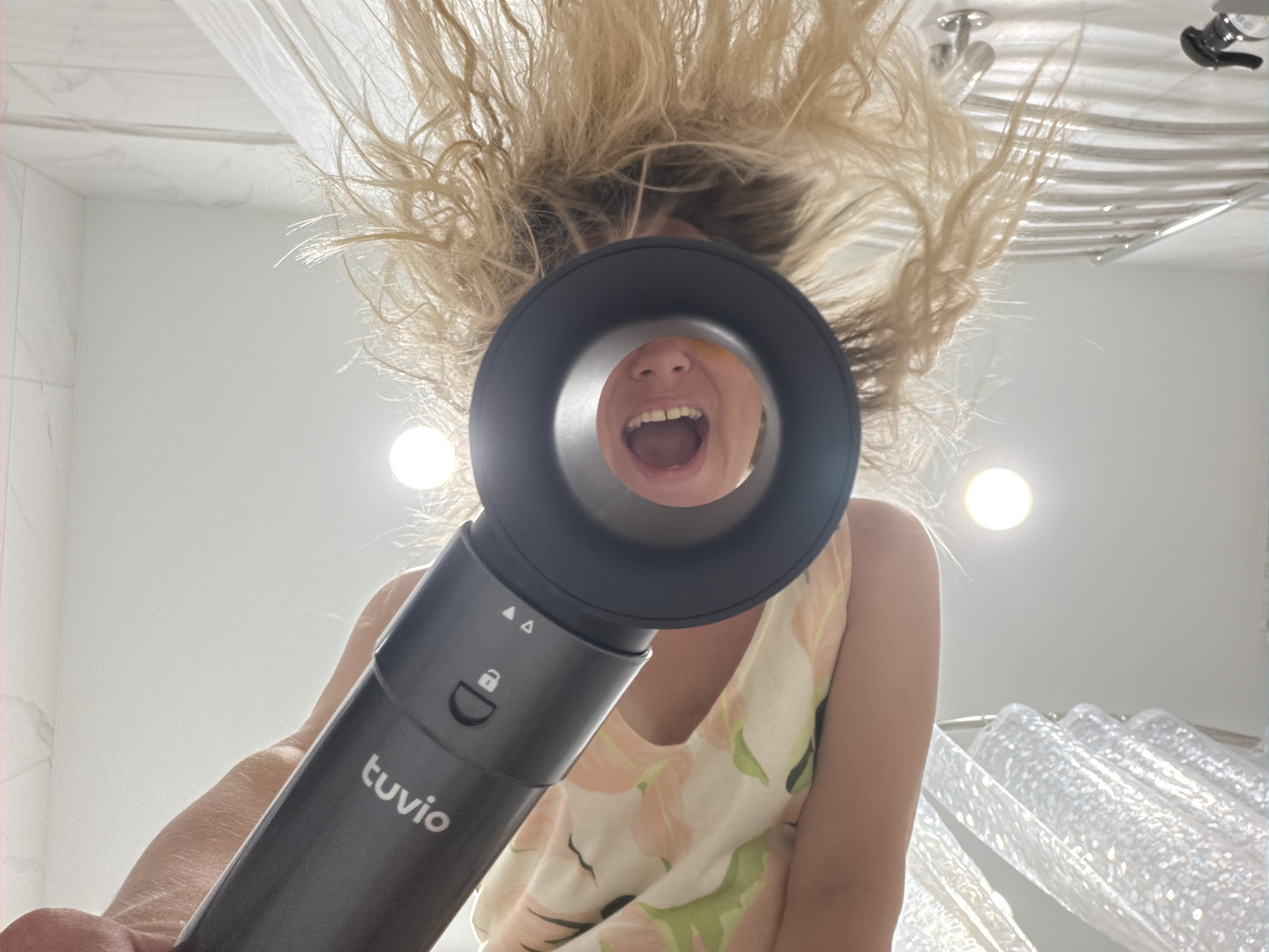 The image shows an extreme low-angle shot, looking upwards into what appears to be a bathroom. A person's hand is holding a dark gray hairdryer with the brand name "tuvio" visible. The nozzle of the hairdryer is directed upwards, and through the opening, a person's mouth is visible, open in a scream or exclamation, with their teeth bared. This individual's blonde, messy hair is also visible, framing the opening of the hairdryer. The background shows a tiled ceiling and a shower curtain. There is no indication of the city of Votkinsk, Russia, in the image.