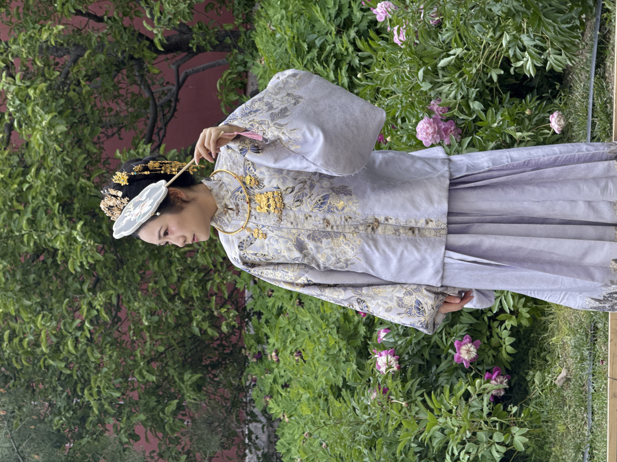 A person in traditional clothing stands among lush greenery in Dongcheng District, China. The individual wears a light purple garment with gold and blue embroidery, along with a golden headpiece and a white fan. The background features a red wall partially obscured by dense green foliage and pink peony flowers. The person is positioned on the edge of a grassy area.