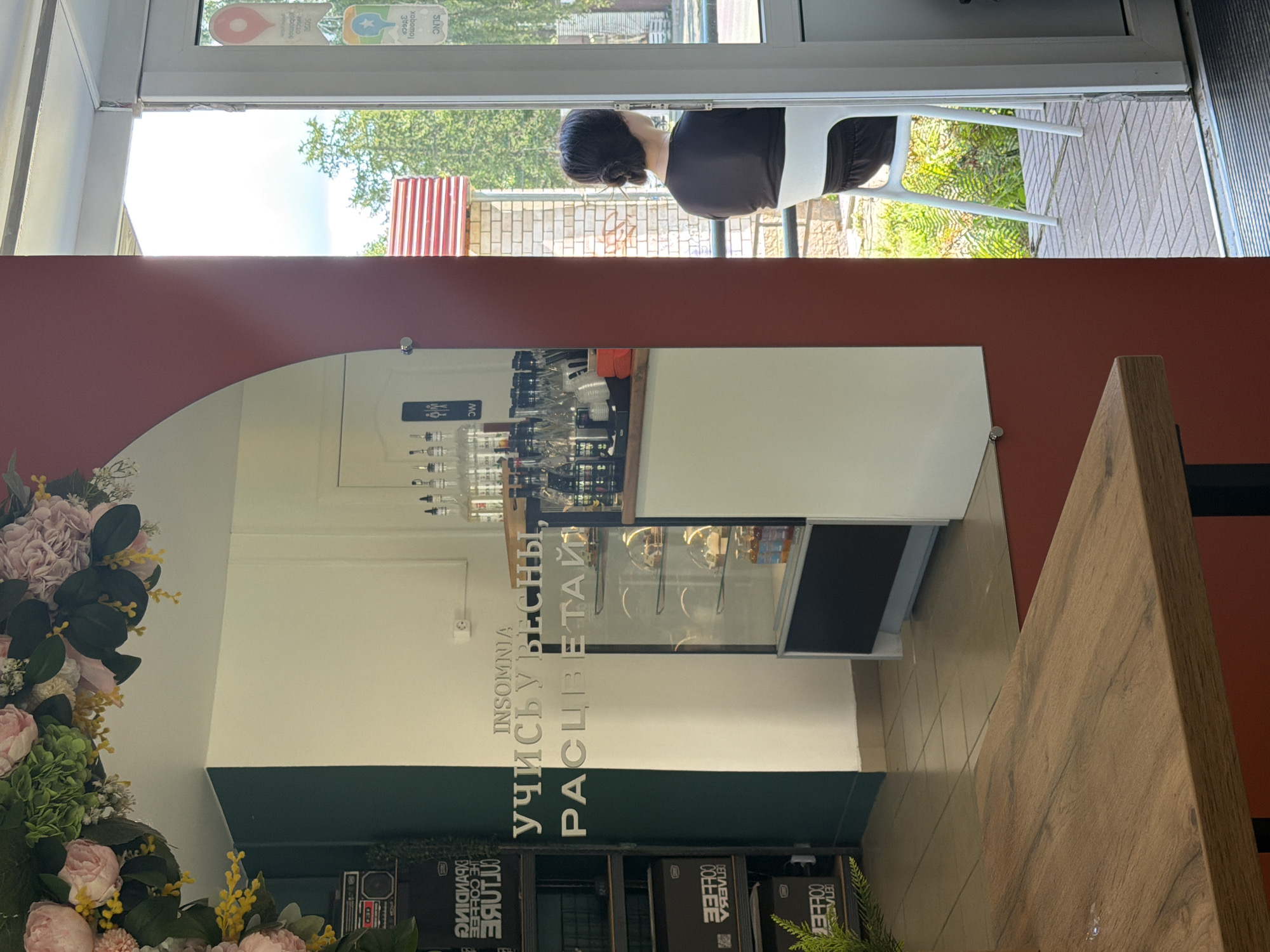 In the city of Chaikovsky, Russia, a cafe interior is depicted with a prominent mirror reflecting a section of the cafe. On the mirrored surface, the words "INSOMNIA УЧИСЬУ ВЕСНЫ, РАСЦВЕТАЙ" are displayed in silver lettering. Behind the mirror, a display case showcases various pastries and bottles. To the left of the mirror, shelves hold boxes and a vintage-looking stereo. Through an open doorway on the right, a woman with her hair in a bun is seated facing away from the camera, looking out at a sunlit outdoor area with greenery. The outdoor area features brickwork and a building facade. The overall lighting suggests daytime.