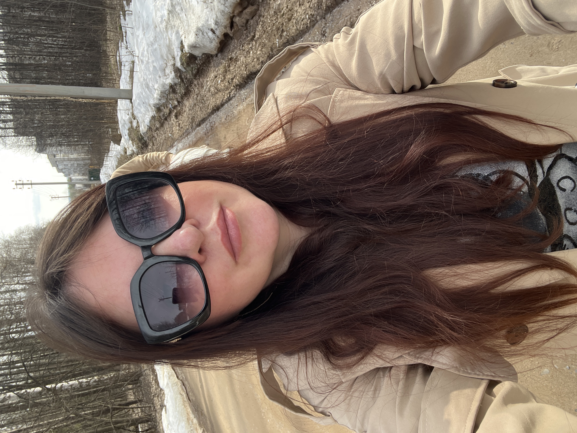 A selfie taken on a road in Noviy, Russia. The photo shows a woman with dark brown hair wearing large, black sunglasses and a beige trench coat. In the background, there is snow and trees. A utility pole is also visible on the left side of the frame.