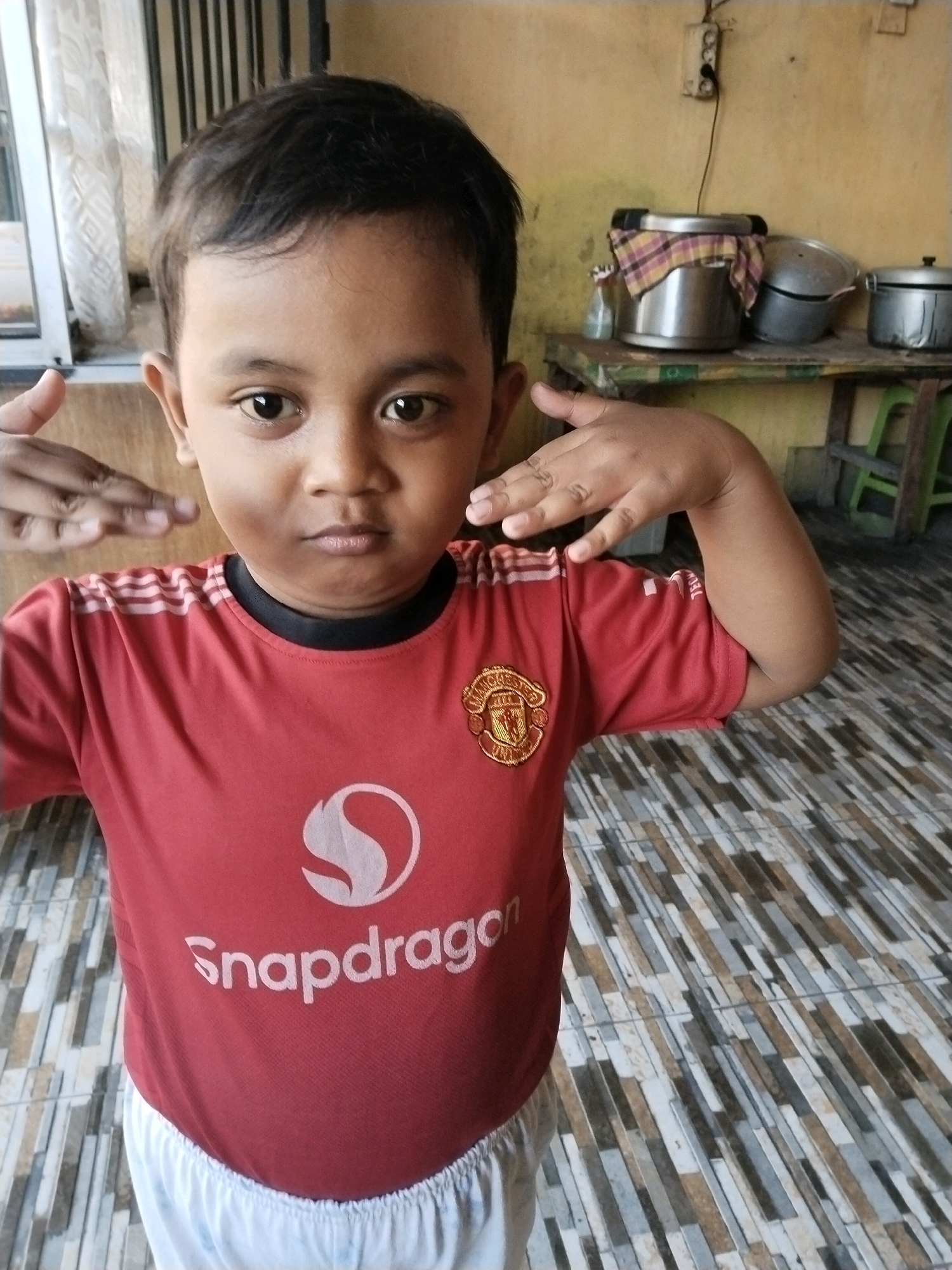 A young East Asian boy stands indoors, facing the camera, wearing a red Manchester United jersey with the Snapdragon logo and white shorts. He has dark hair and is holding his hands up near his temples, with his fingers splayed. The background shows a yellow wall, some metal pots on a shelf, and a tiled floor. The setting appears to be a humble residence in Pepelegi, Indonesia, likely a kitchen or living area, suggesting a casual, everyday moment.