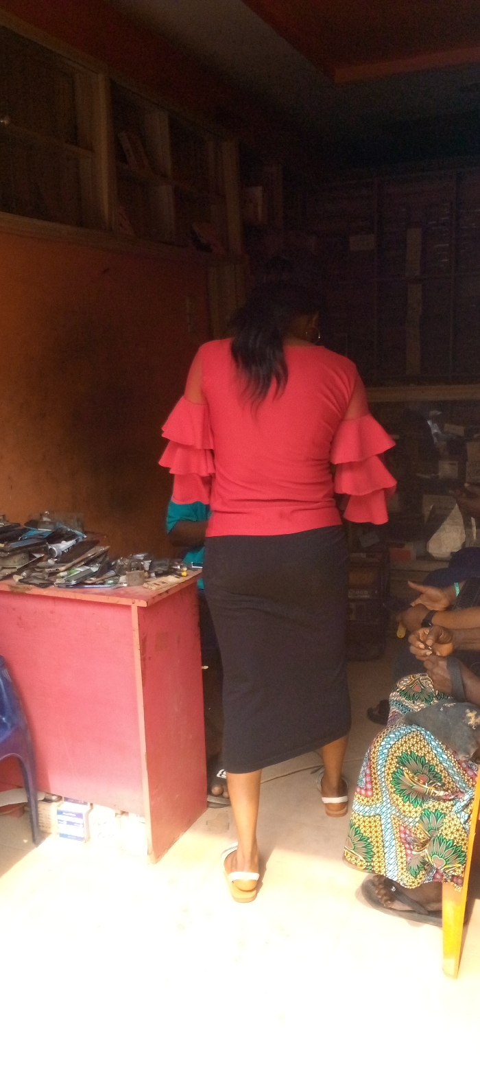 The image depicts an indoor scene within a commercial establishment in Nnewi, Nigeria, likely an electronics repair shop or a vendor of mobile phone components.
In the foreground, a female subject stands with her back to the camera, positioned centrally. She wears a red short-sleeved top with triple ruffles on each sleeve, a black midi skirt, and light-colored slip-on sandals. Her dark hair is pulled back into a ponytail.
To the left of the central subject, a reddish-brown wooden counter holds numerous electronic devices, primarily mobile phones and their disassembled parts, including screens, casings, and circuit boards, arranged in piles. A partially visible person wearing a blue and red top stands behind or next to this counter, obscured by the central subject. A blue plastic chair is visible in the lower-left corner, next to the counter.
To the right of the central subject, a seated individual is partially visible, showing their lower torso and legs. This person wears a colorful patterned skirt (likely Ankara fabric) and dark flip-flops. Their hands are clasped in their lap. A wooden chair leg is visible near their feet.
The environment features reddish-brown or orange-painted walls. In the upper-left background, a wooden shelving unit or window frame contains indistinct items, possibly books or files. The floor is light-colored, with a bright area in the foreground indicating a light source, likely a doorway or window, ahead of the central subject. The rear of the establishment appears darker. No discernible interactions between the subjects are depicted.