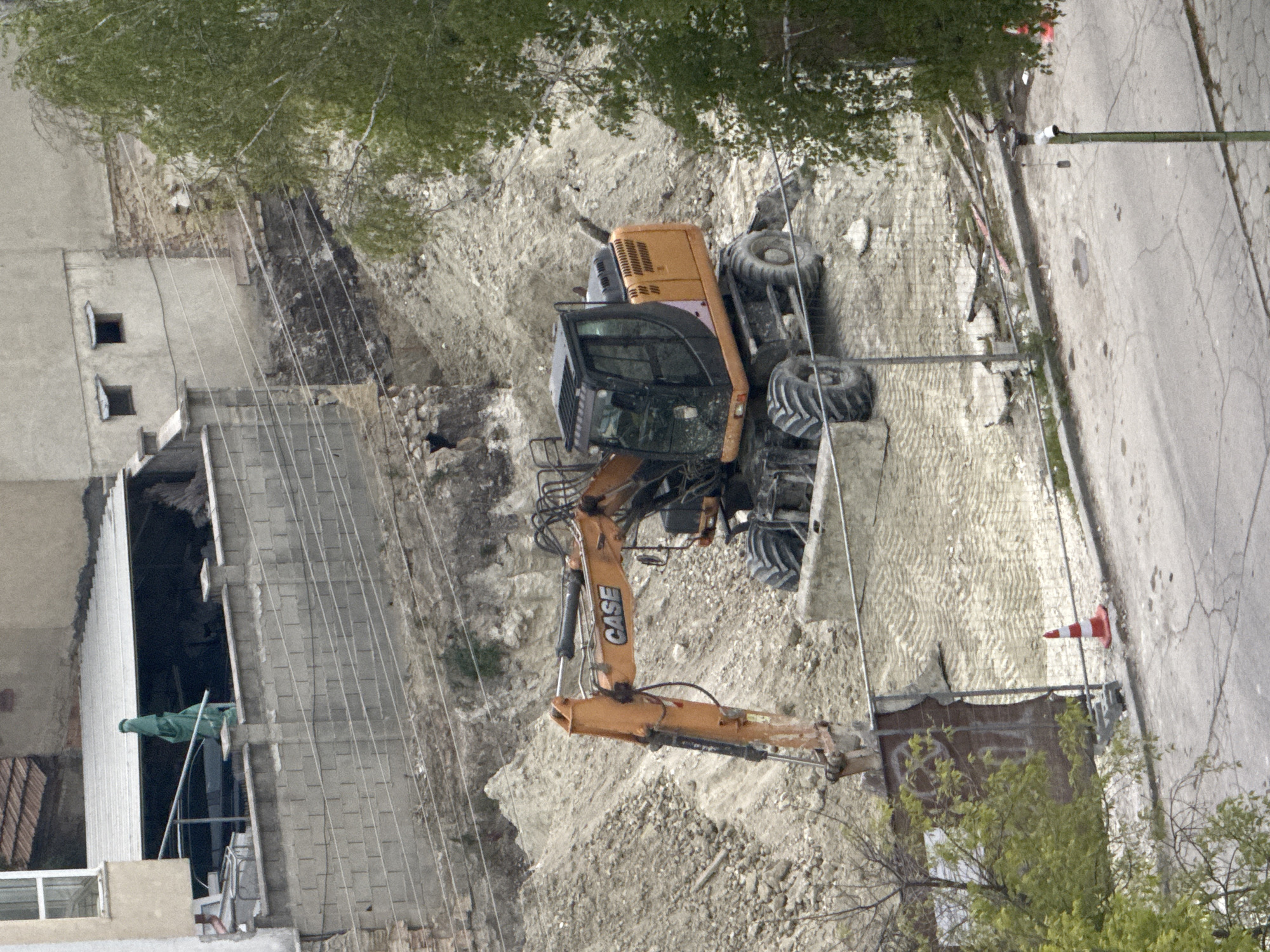 This media file, oriented sideways, captures a construction or demolition site in Varna, Bulgaria, under diffused daylight conditions.
The central subject is a large orange and black wheeled excavator, prominently displaying the "CASE" brand name on its boom. The excavator is positioned on an uneven, excavated area of sandy and rocky earth, which appears to be a cleared ground where work has taken place or is ongoing.
In the background, on what would be the left side if the image were correctly oriented, stand several buildings. Some appear to be under construction or partially demolished, with exposed concrete block walls and scaffolding-like structures. Multiple utility lines or cables stretch across the scene, connecting to structures or poles. Green foliage from trees is visible, with branches extending into the frame.
Towards the right side of the image, a paved road with visible cracks runs along the edge of the construction area, separated by a curb. A red and white traffic cone stands on the side of the road. More green trees and sparse vegetation are also seen near the road.
No people are visible in the image. The scene suggests an active urban development or renovation project. The lighting indicates it is daytime, possibly overcast or with indirect sunlight.