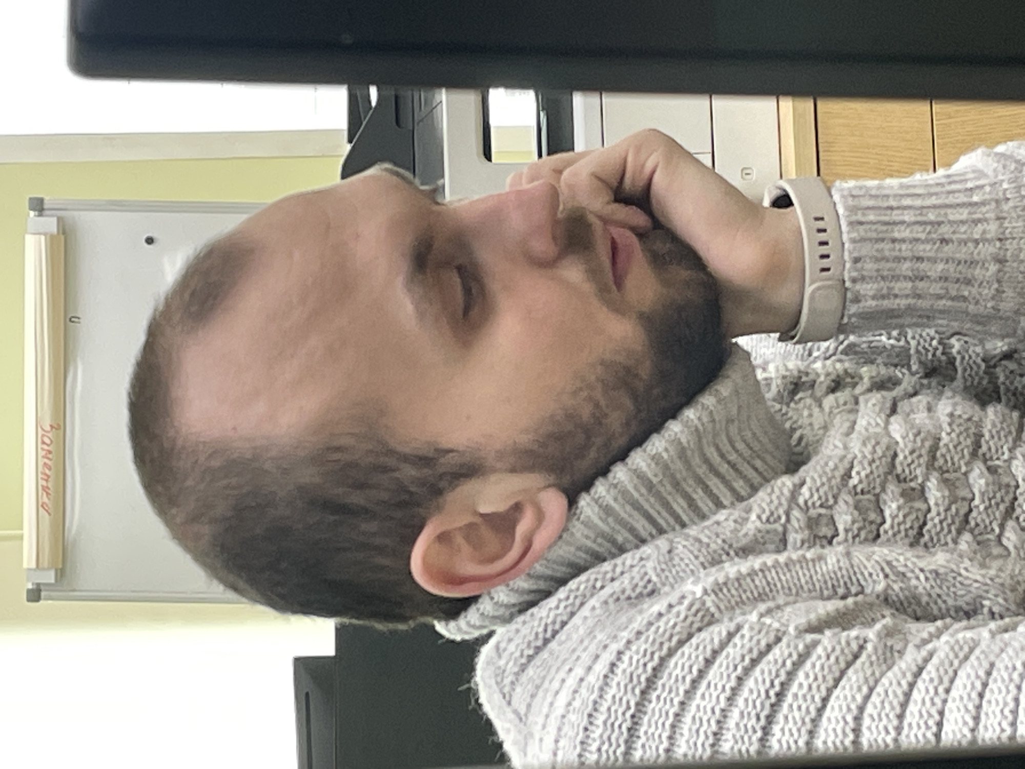 A fair-skinned man with short, thinning brown hair and a beard is sleeping, resting his head on his hand. He is wearing a gray turtleneck sweater and a light-colored smart watch. The setting appears to be an office or workspace, with a monitor visible in front of him. A whiteboard with Russian text ("Заметки", meaning "Notes") is partly visible in the background, which may suggest this is Krasnoyarsk, Russia.