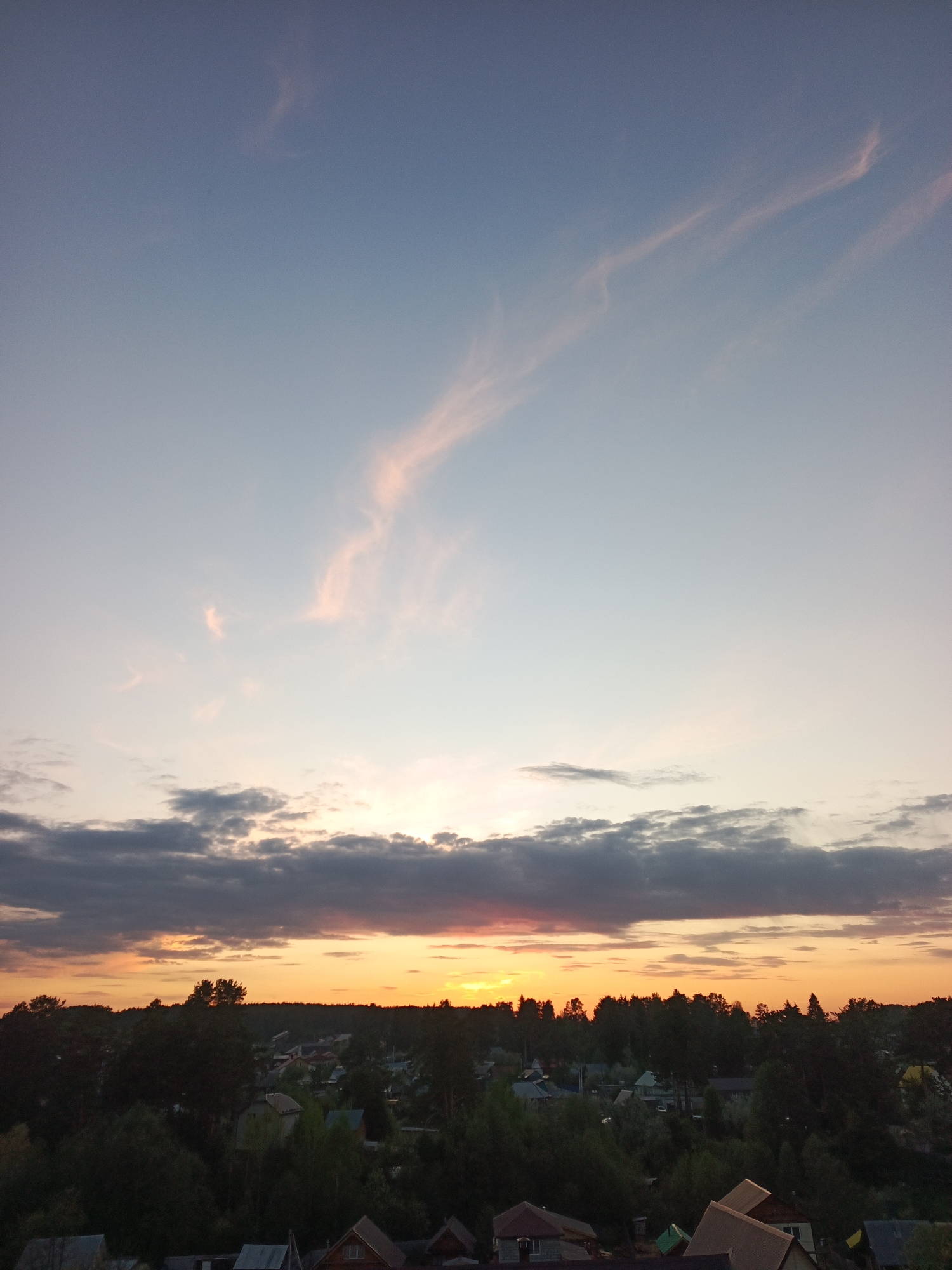 The image shows a sunset in Chaikovsky, Russia. The sky is a mix of blue and orange, with pink streaks in the upper part. A band of gray clouds stretches across the horizon, below which the sunset casts an orange glow. Below the horizon, trees and rooftops of houses can be seen. The scene appears to be captured from an elevated perspective.