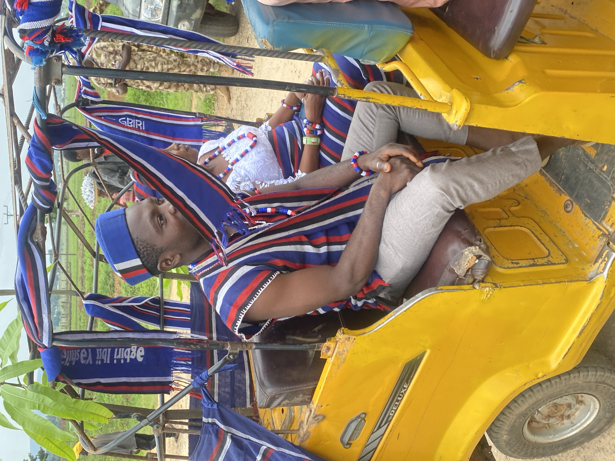 Two individuals are inside a yellow tricycle. The person in the foreground wears a blue hat and a striped shirt of red, white, and blue. They are wearing gray pants, with their hands clasped together. The individual behind them has on white clothing and beaded necklaces and bracelets of white, red, and blue. The tricycle is decorated with striped material of blue, white, and red. The words "agbiri ibi ya state" can be read on one of the decorative pieces, written in reverse. The location could be Maigizo, Nigeria.