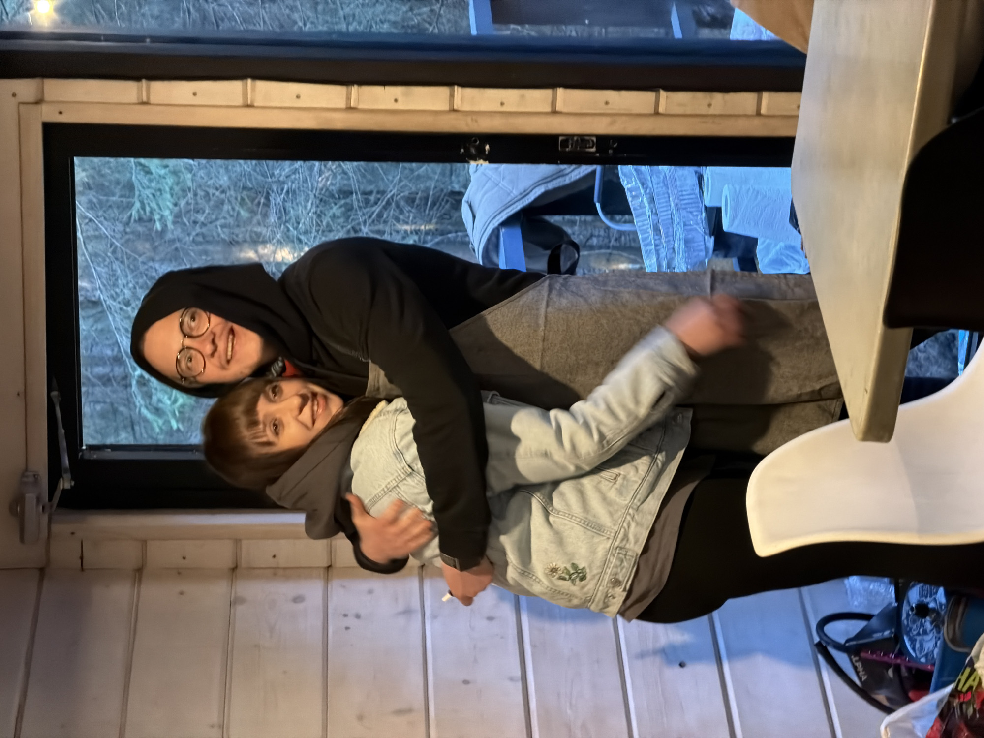 Two people are standing inside a wooden-paneled building with a window. The man is wearing a black hoodie and glasses and is embracing the woman from behind. The woman is wearing a light denim jacket over a grey hoodie and black leggings. The window looks out onto a forest with bare trees. A table and white chair are partially visible in the foreground. There is no evidence to indicate that this image was taken in Vsevolozhsk, Russia.