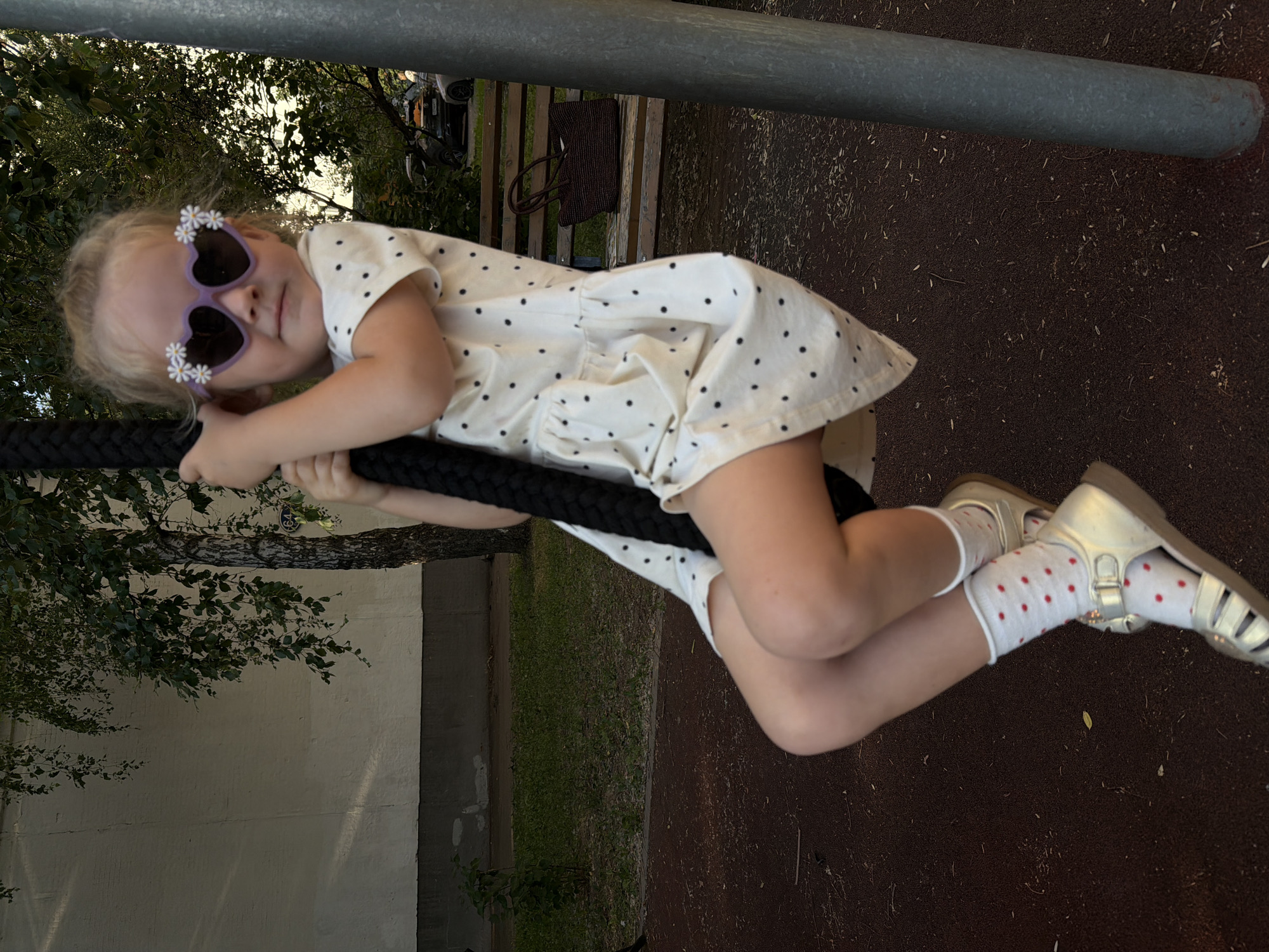 This image shows a young girl on a playground in Khimki, Russia. She is wearing a white dress with black polka dots, white socks with red polka dots, and gold sandals. She is also wearing heart-shaped sunglasses with small white flowers around the lenses. The girl is holding onto a thick black rope, likely part of a swing or climbing structure, and appears to be in motion. Her legs are bent, and she is looking towards the camera with a slight smile.
In the background, there is a wooden bench with a dark brown bag on it. Behind the bench, there are trees and a building. The ground of the playground appears to be a dark, possibly rubberized, surface. The lighting suggests it is daytime, likely late afternoon given the shadows. The overall mood is playful and candid. No visible text is present in the image.