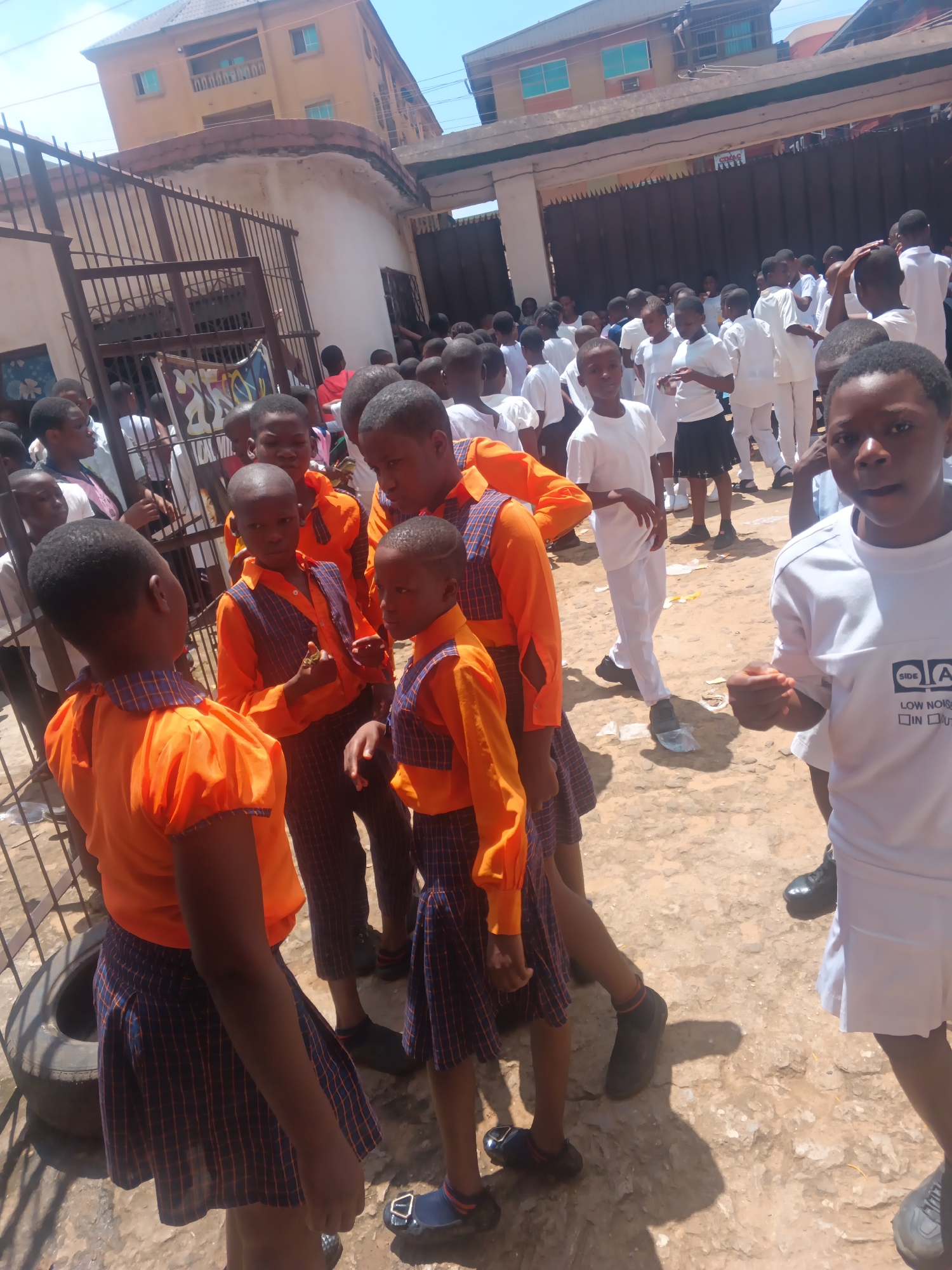 In Onitsha, Nigeria, a group of schoolchildren are gathered outdoors on a dirt ground, likely within their school premises. Many of the children are wearing school uniforms. Several children in the foreground are wearing bright orange shirts, some with blue and orange plaid vests and matching plaid skirts or trousers. Others are wearing white shirts and trousers or skirts. The children appear to be of various ages, and their interactions suggest they are engaged in a school activity or gathering. In the background, a multi-story building, possibly a school building, is visible, along with a metal fence and gate. The overall environment appears to be an urban setting under a clear sky.