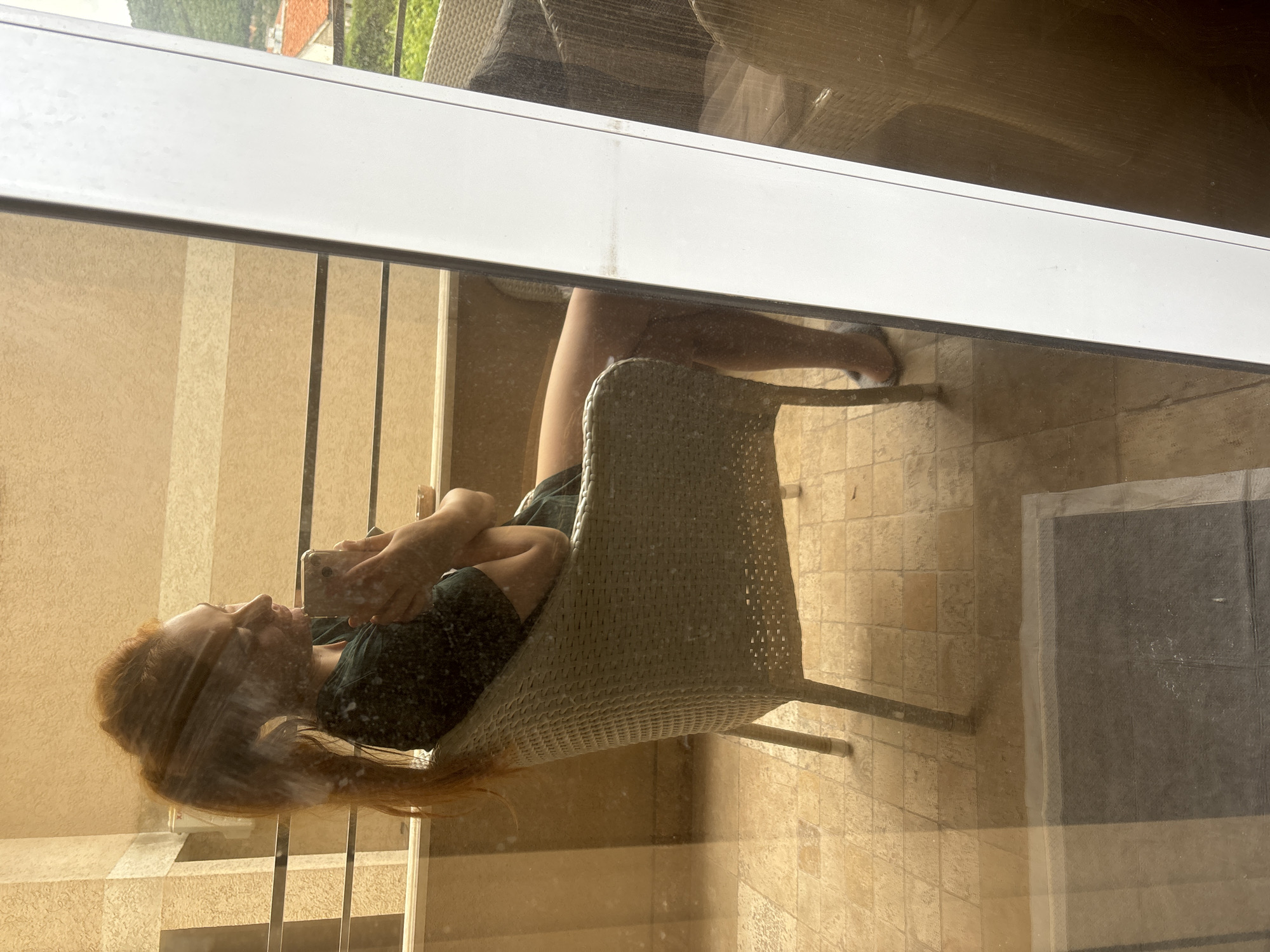 The image shows a person with long red hair sitting in a wicker chair on a balcony, seen through a glass door. The person is holding a phone and appears to be looking at it. The balcony has a beige tiled floor and a railing. Reflected in the glass door is another part of the balcony with a view of a building with an orange roof and some greenery. Based on the architectural style and landscape, the location is likely Budva, Montenegro.