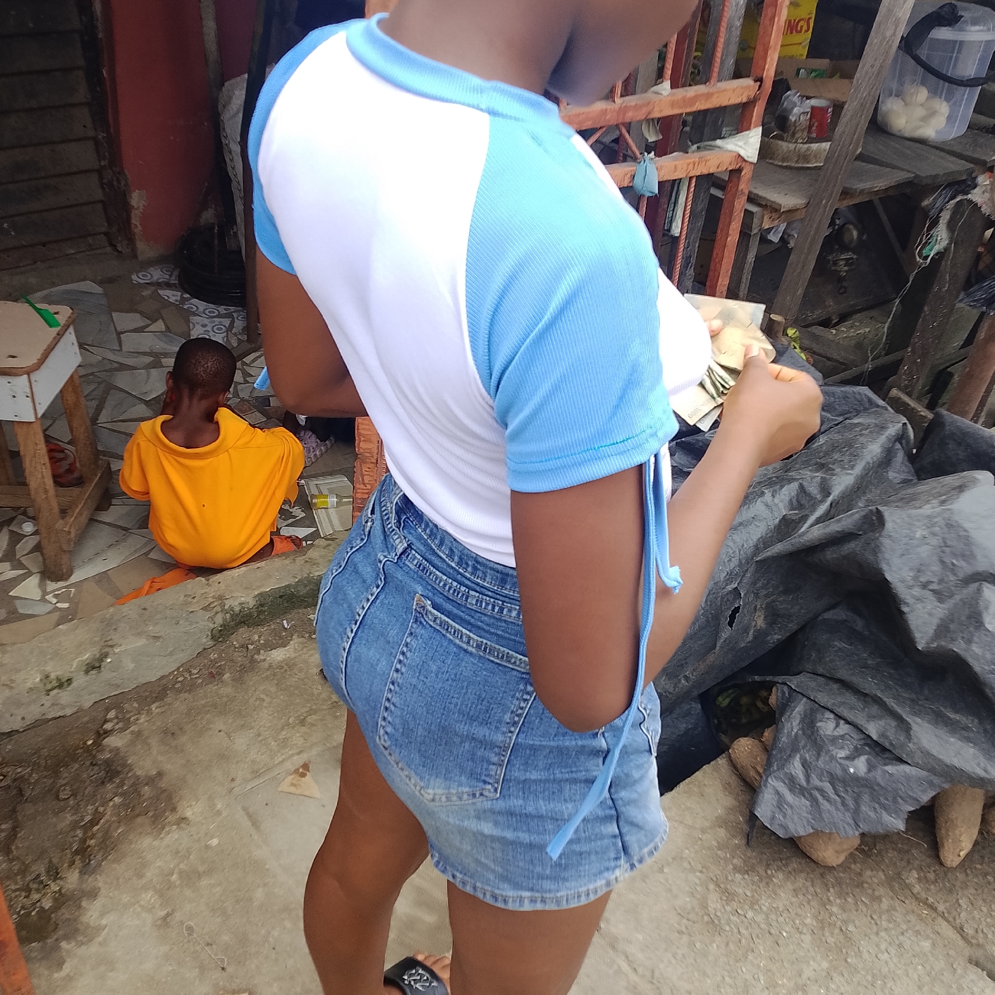 This image depicts a street scene in Ado, Nigeria. In the foreground, a person is seen from the side wearing a white t-shirt with light blue sleeves and a denim skirt. Their hands are holding some banknotes. To the left, a child in an orange t-shirt and orange pants is sitting on the ground, facing away from the camera. The environment shows a rough, unpaved ground surface with some scattered debris. Behind the subjects, there is a makeshift stall or structure with wooden planks and metal bars, displaying various items like plastic containers, cans, and what appear to be root vegetables covered by a black tarp. The overall impression is that of a typical informal market or roadside area.