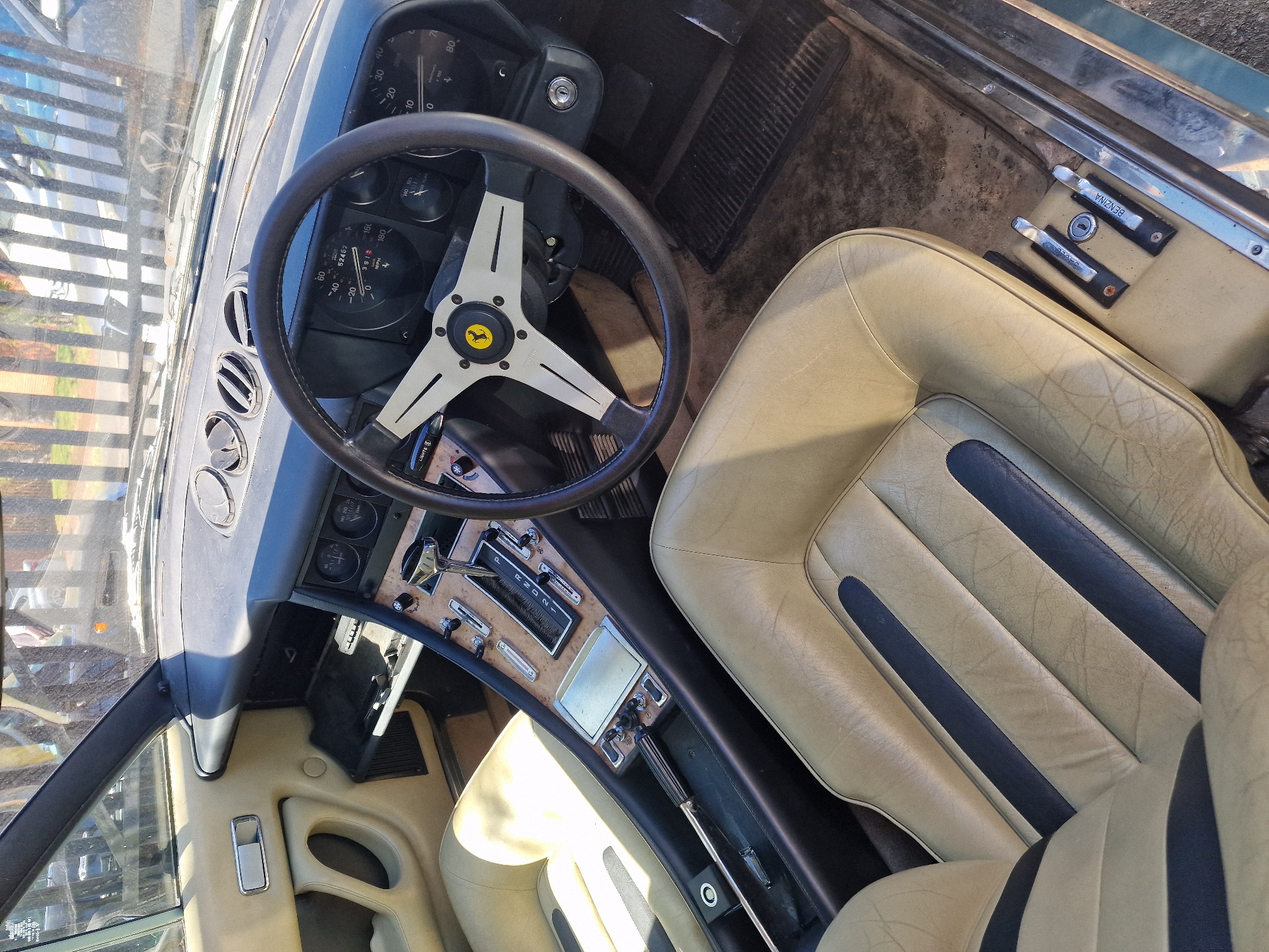 This close-up photograph captures the interior of a classic car, likely a Ferrari given the emblem on the steering wheel, taken from an elevated perspective looking down into the driver's side. The scene is set outdoors in South Tyneside, United Kingdom, under bright, sunny daylight.
The primary subjects are the car's dashboard, steering wheel, seats, and central console. The steering wheel, featuring a black rim and silver spokes, prominently displays the yellow Ferrari logo with a prancing horse. Behind it, the dashboard houses various analog gauges, including a speedometer showing a reading of "52452" miles and marked with "MPH" up to "180," and a tachometer marked "x 1000" up to "80." A switch labeled "LIGHTS" is visible below the steering wheel.
The seats are upholstered in a light cream or beige leather with distinctive dark blue vertical stripes, showing some creasing and wear consistent with age. The central console, partially covered in a wood-grain trim, features a chrome-handled automatic gear selector with positions "P R N D 2 1" clearly visible. Adjacent to the passenger seat, two pull handles are present, one labeled "BENZINA" (Italian for fuel/gasoline) and another, likely for the hood or trunk release, partially visible with "APRI" (Italian for open). The interior appears well-maintained for its age. No people are visible in the image, suggesting the car is parked.